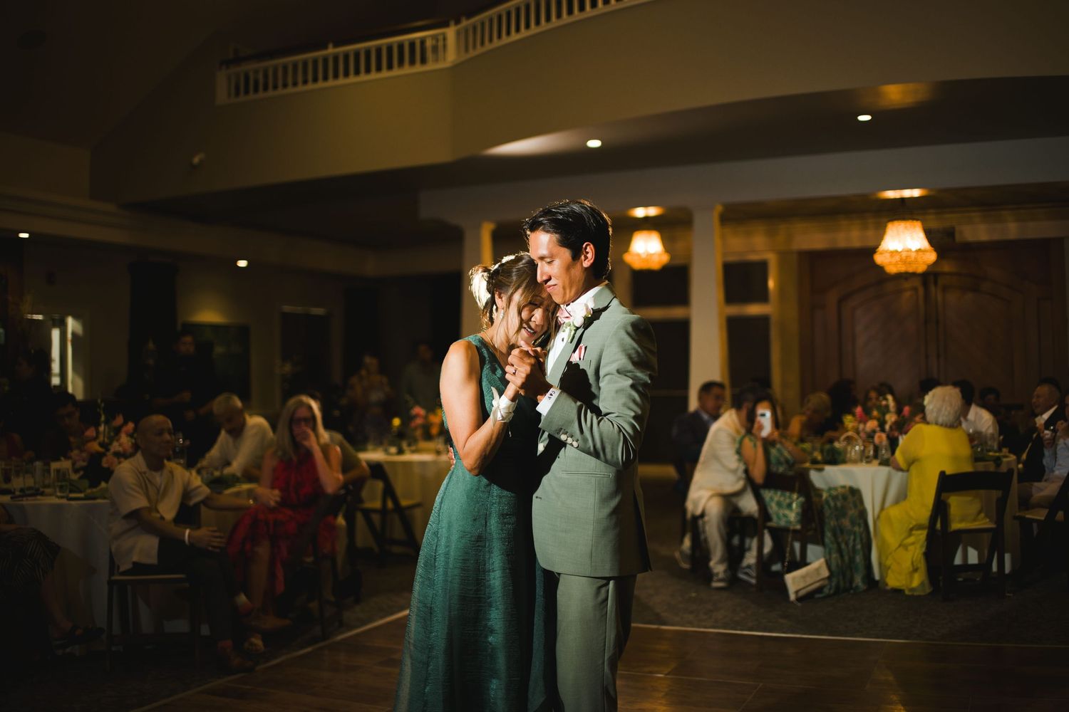 Couple shares first dance at elegant wedding reception with guests seated at candlelit tables in warm ambient lighting.