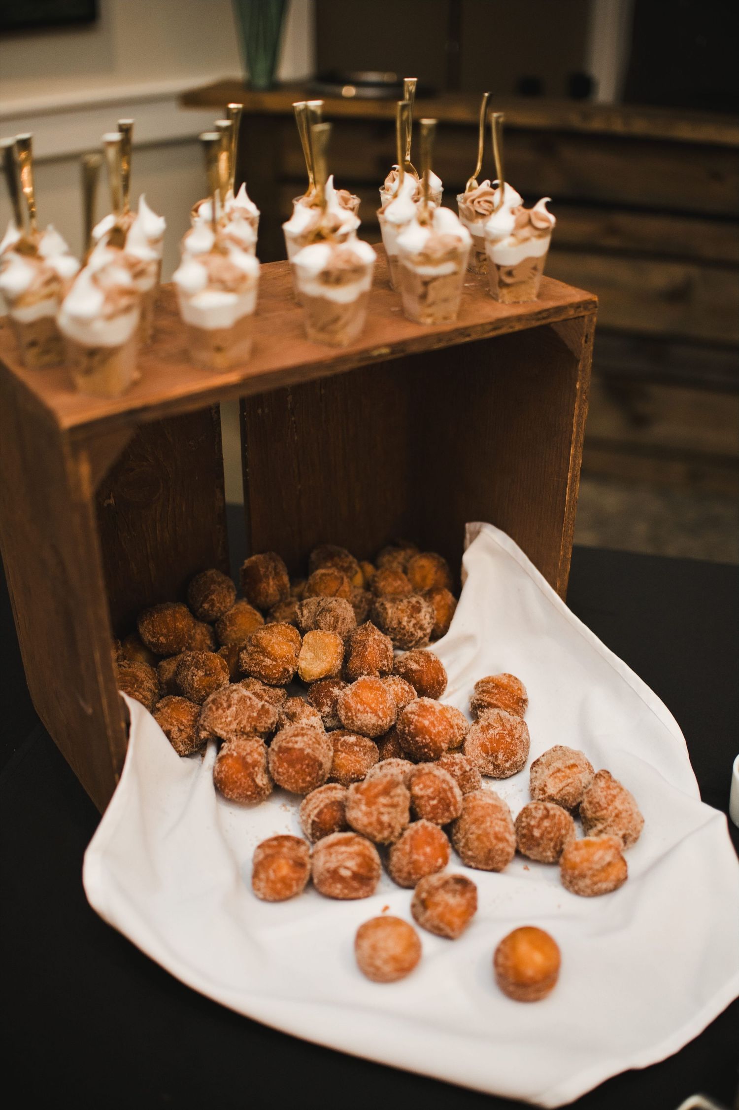 Elegant dessert display with sugared donut holes and cream-topped mini dessert cups on a rustic wooden crate.