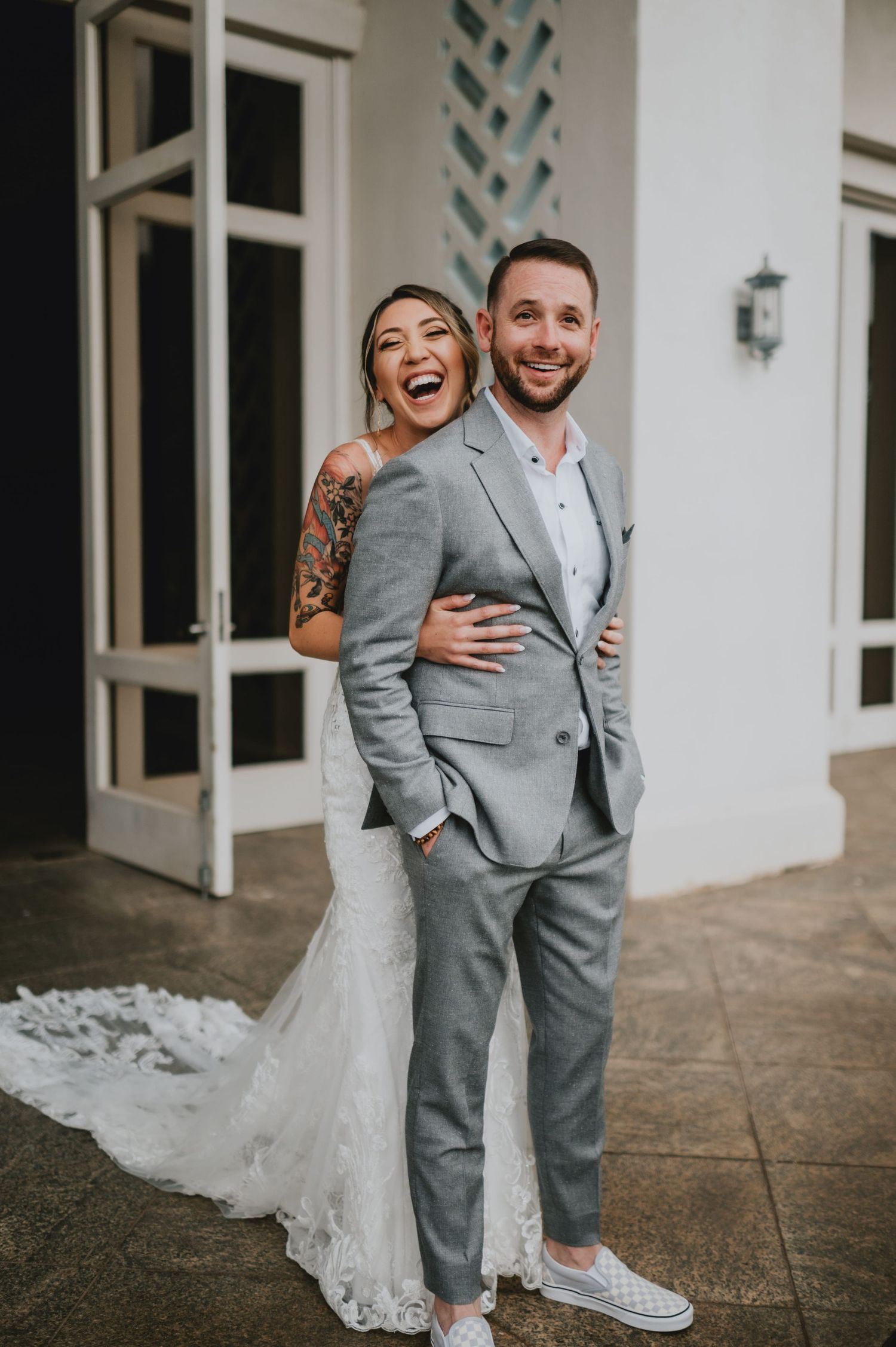 Laughing bride in lace gown with tattoos embraces smiling groom in gray suit and white sneakers outside a white building.