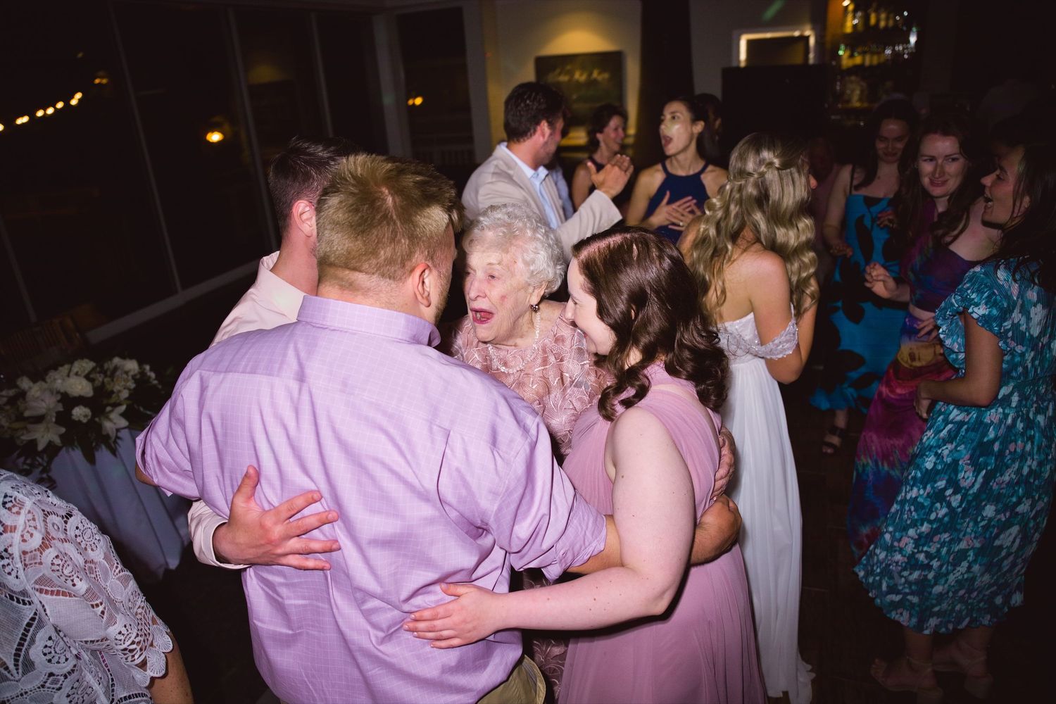 Guests dancing joyfully at a wedding reception, with an elderly woman in a floral dress laughing among the crowd.