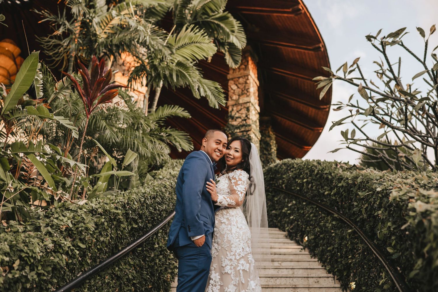 A couple in wedding attire embraces on garden steps surrounded by tropical greenery and a wooden structure.