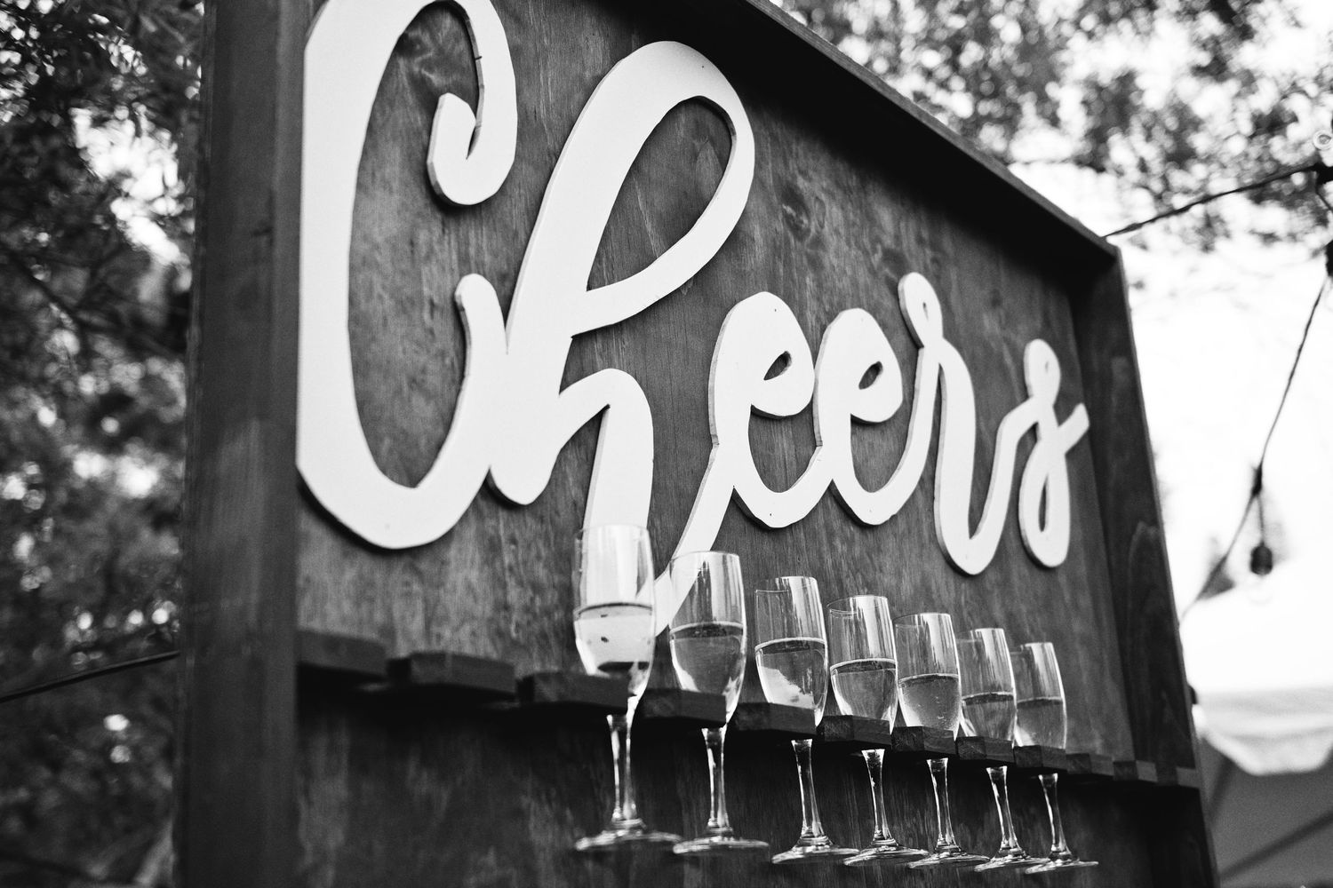 Black and white photo of a rustic wooden Cheers sign with a row of champagne glasses at an outdoor event.