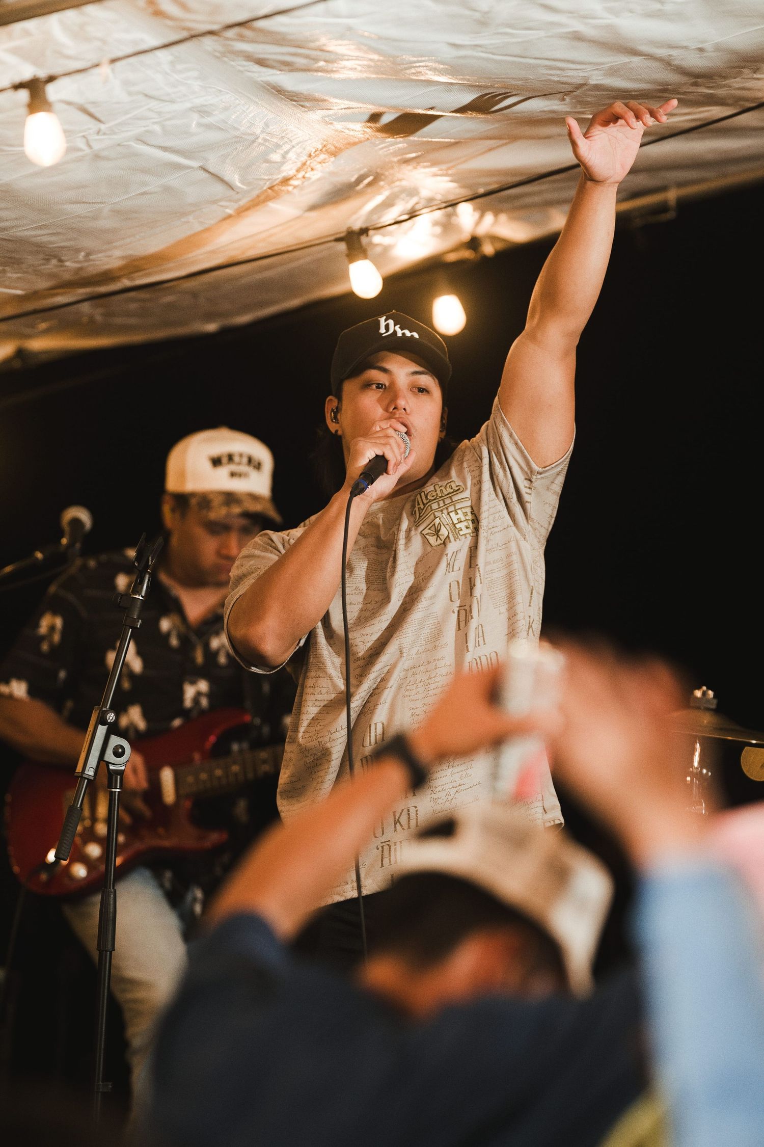 Singer performing on stage with one arm raised, holding microphone, with guitarist in background at live music event.