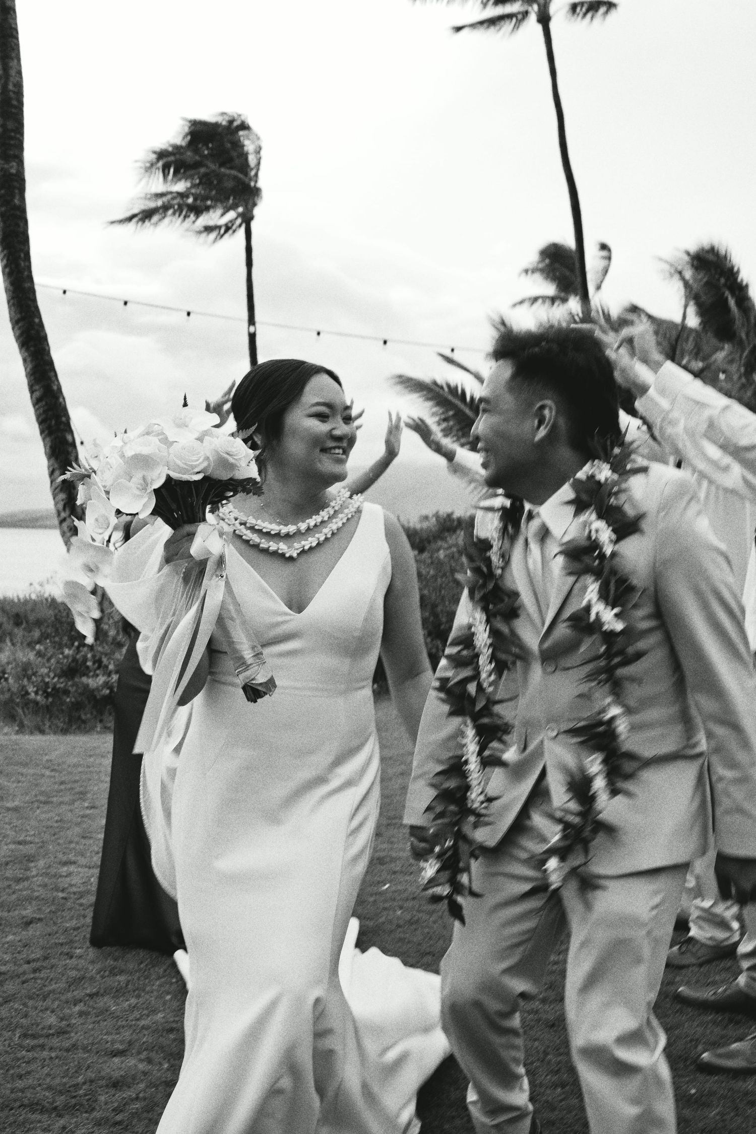 Black and white photo of a smiling bride and groom in leis walking together at an outdoor Hawaiian beach wedding.