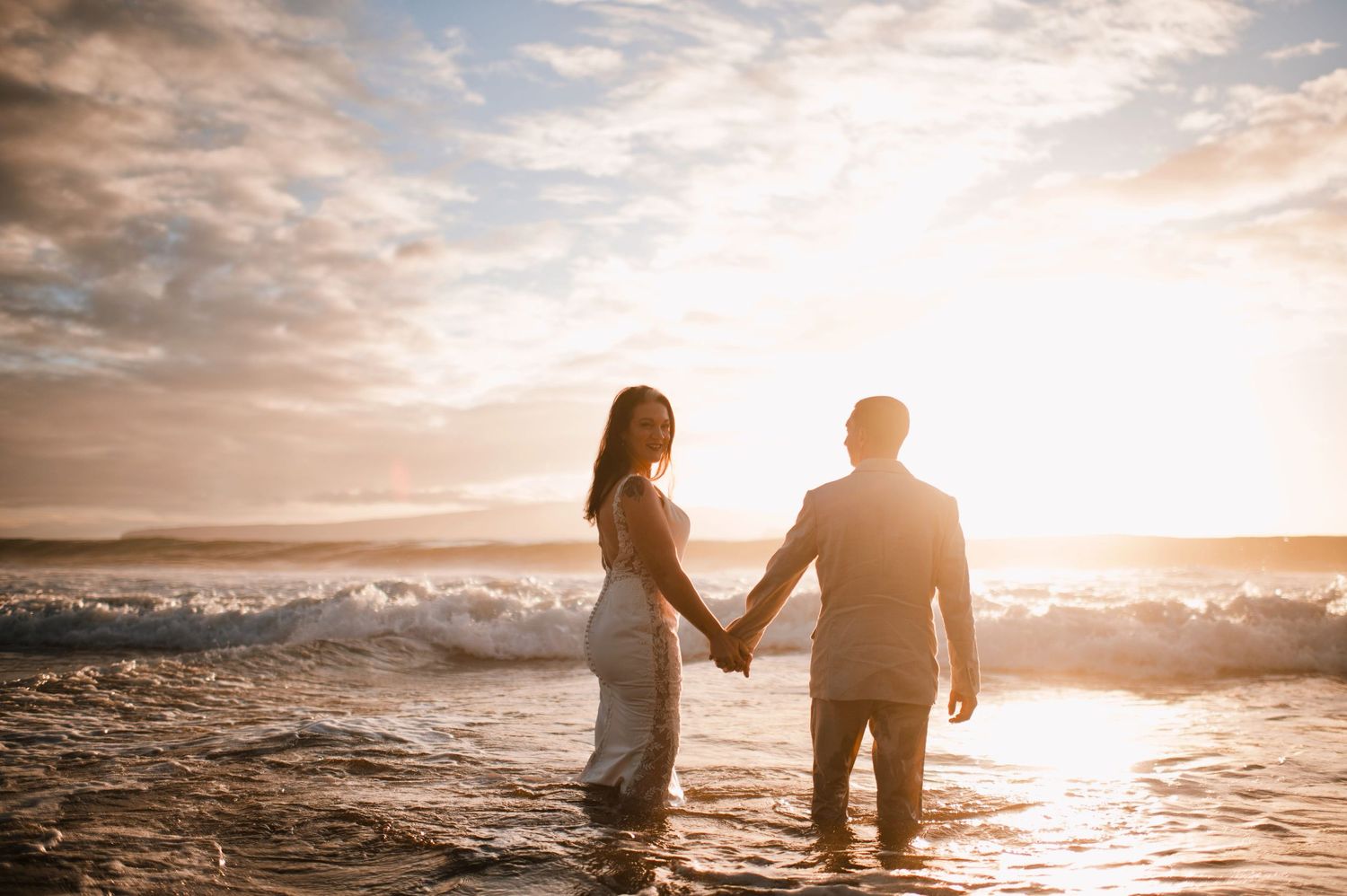 Couple holding hands at sunset on a beach with golden waves crashing behind them.