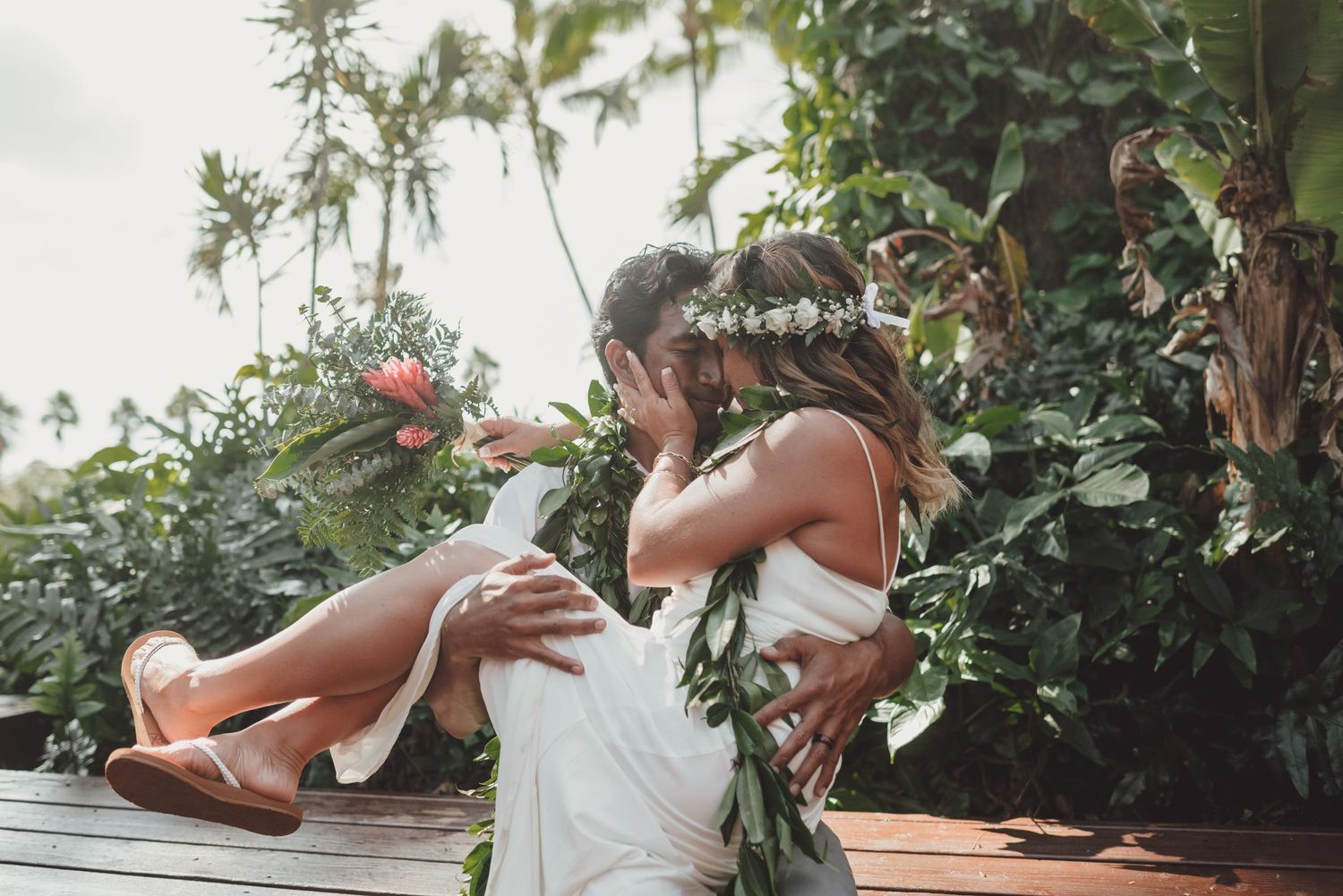 Couple sharing a romantic kiss outdoors, bride in white dress with floral crown, groom lifting her amid lush tropical greenery.