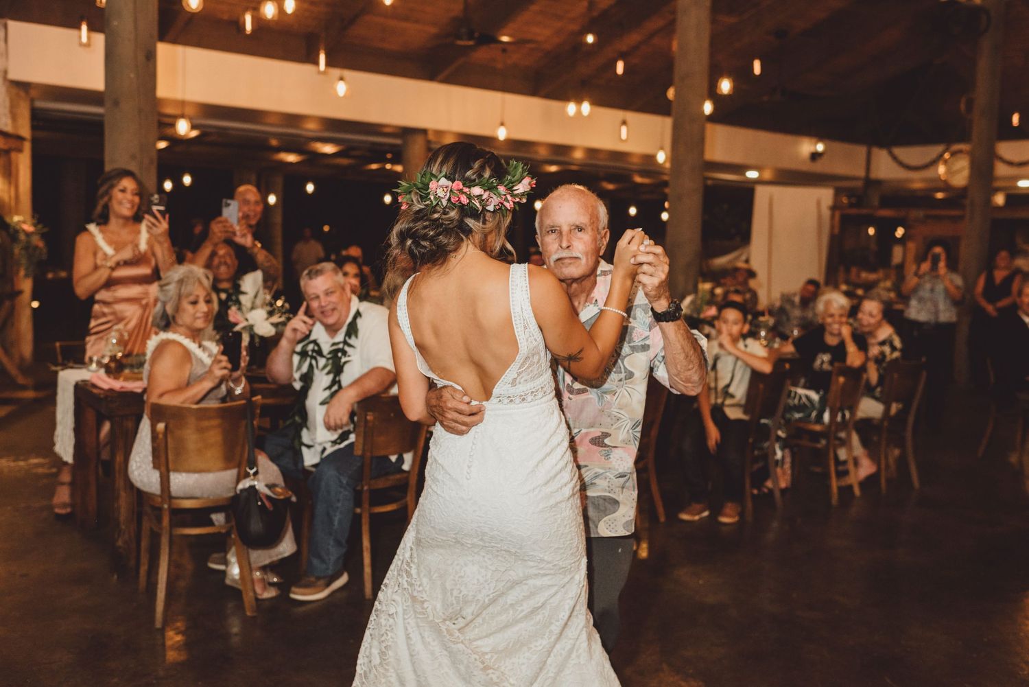 Bride in white gown dances with older man at rustic wedding reception while guests watch and celebrate.