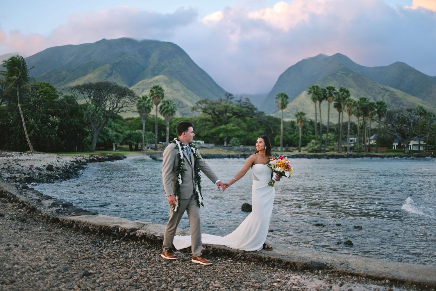 Newlyweds hold hands on rocky shoreline with tropical mountains and palm trees backdrop.