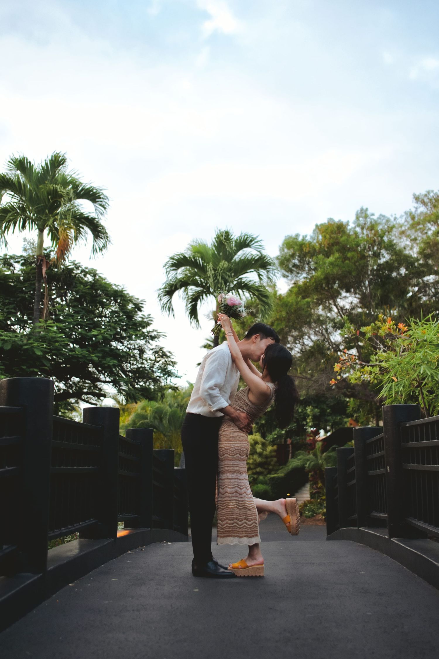 Romantic couple embracing and kissing on a bridge surrounded by tropical trees under a blue sky.
