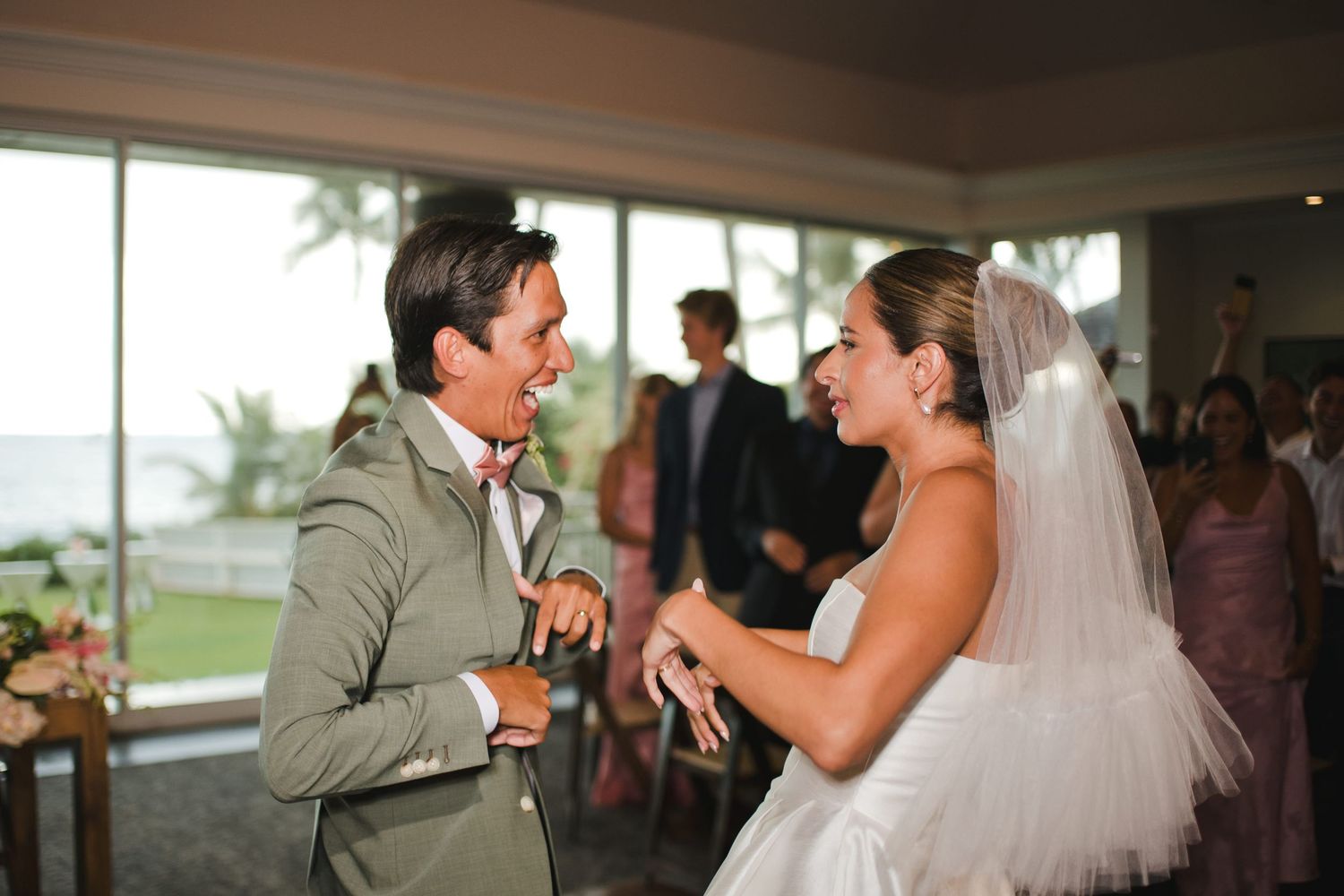 Bride and groom laughing joyfully during wedding reception with ocean view backdrop.