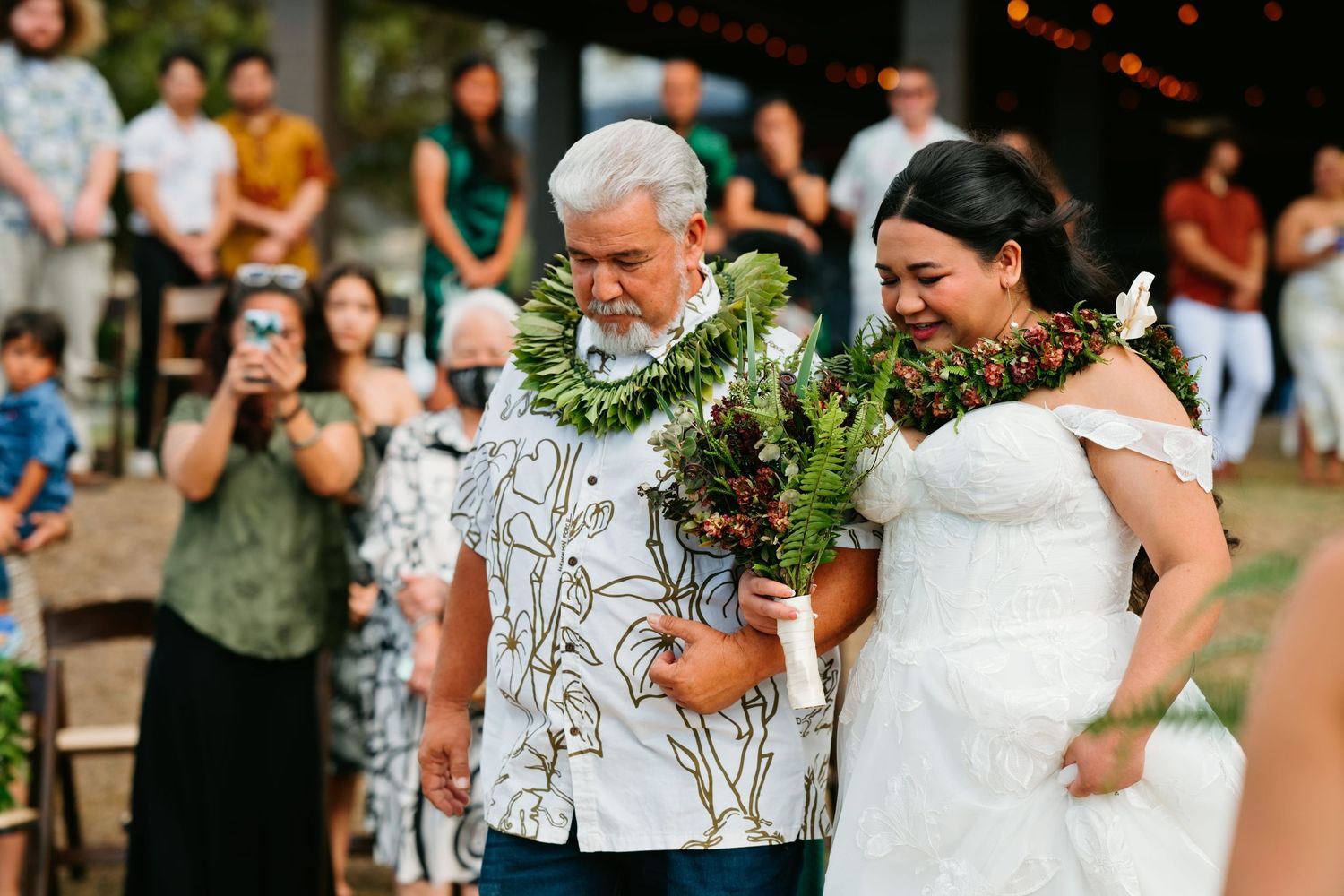 Bride in white off-shoulder gown walks down aisle with older man in Hawaiian shirt, both wearing floral leis.