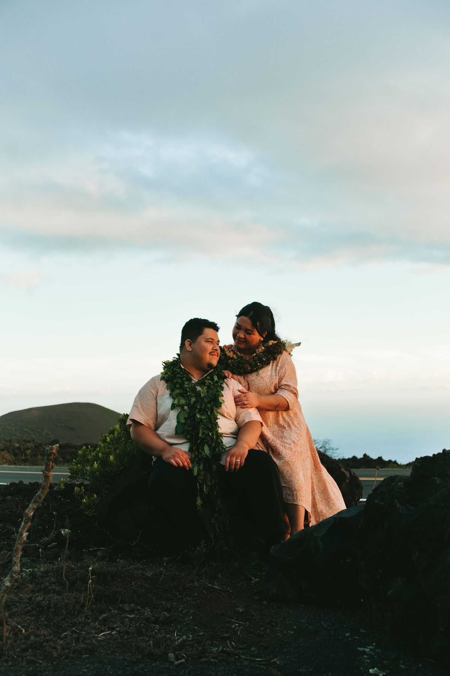 Couple wearing Hawaiian lei garlands sitting on volcanic rocks at sunset with ocean and mountains in background.