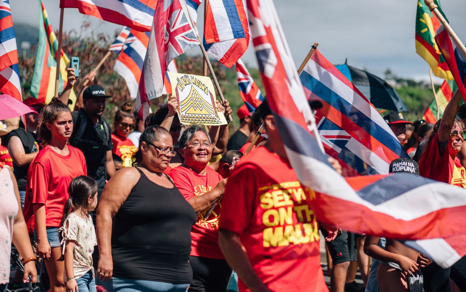 People in red shirts wave Hawaiian flags during organized protest event.
