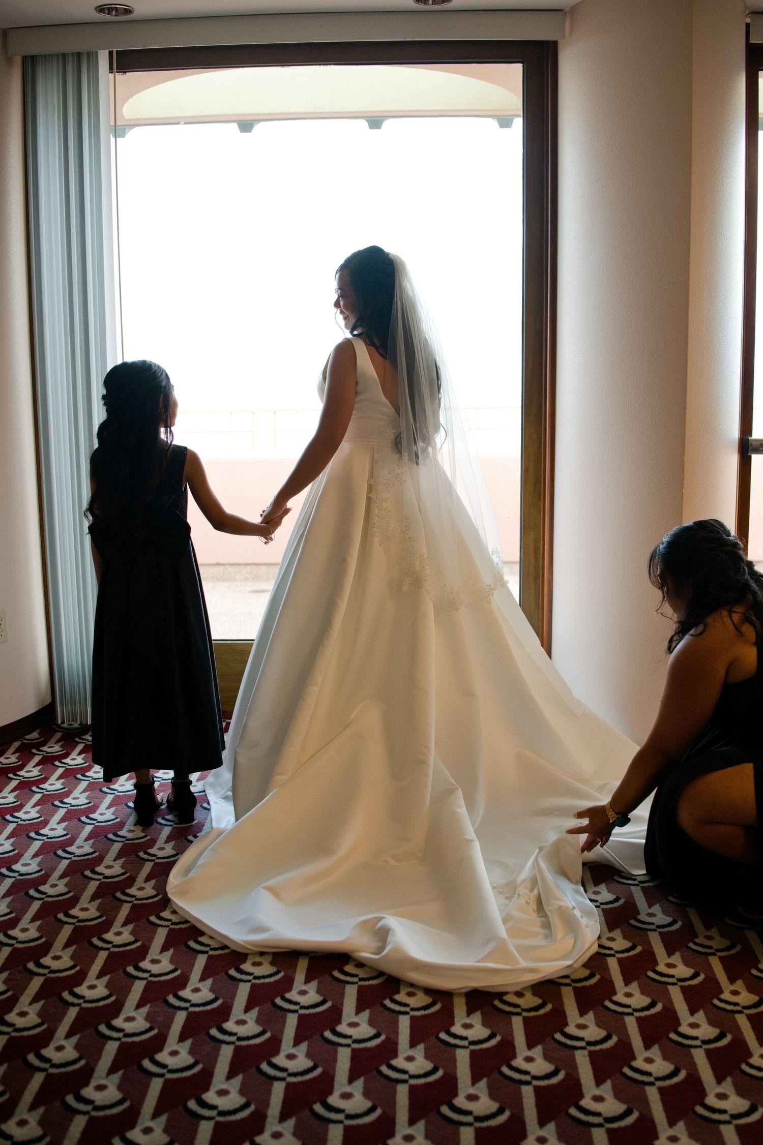 Bride in white gown holds flower girl's hand by window as bridesmaid adjusts train on patterned carpet.