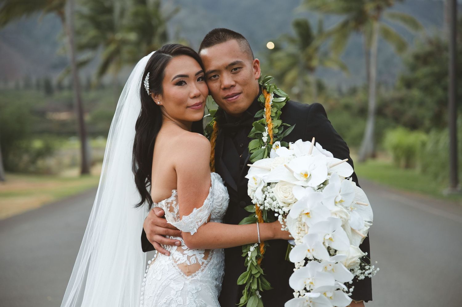 Bride and groom posing together on a scenic road, bride in lace gown with veil, groom holding white orchid bouquet.