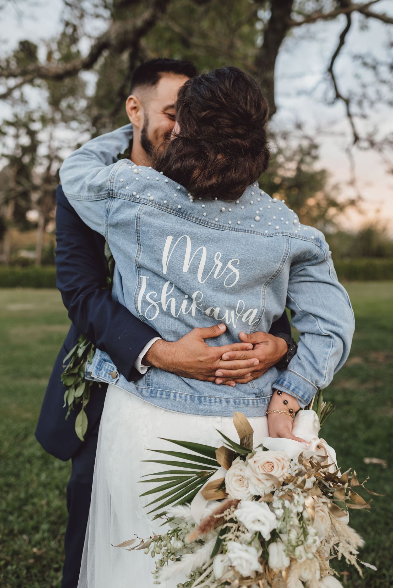 Bride in custom Mrs. Ishikawa denim jacket embracing groom outdoors, holding a bouquet of white and blush flowers.