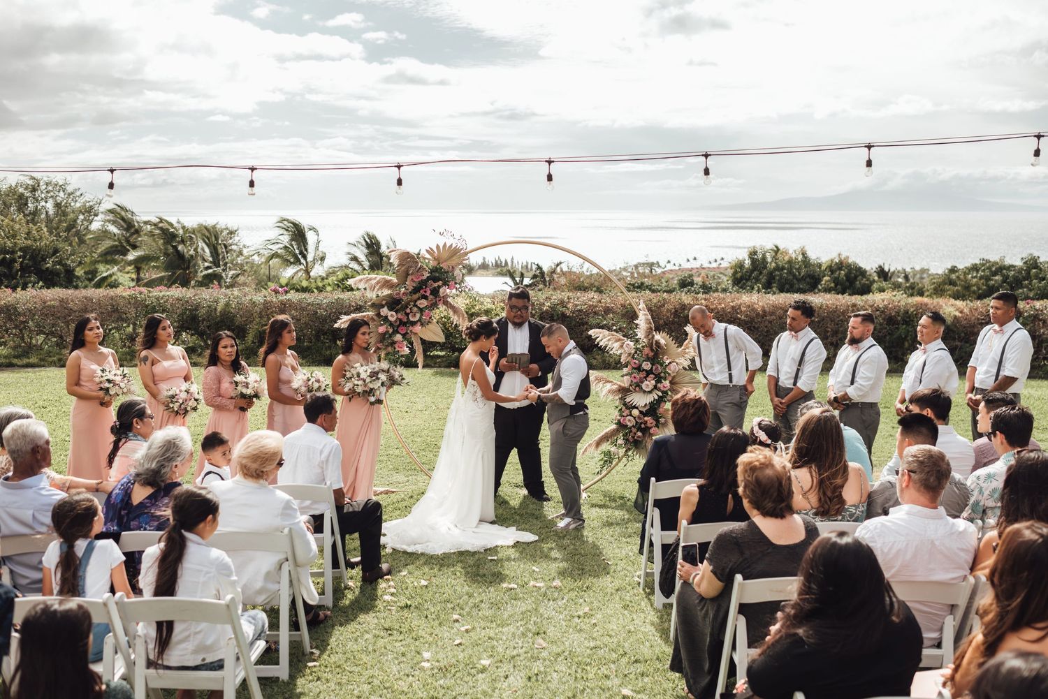 Outdoor wedding ceremony on green lawn with bridesmaids in pink, groomsmen, and guests seated in white chairs under string lights.