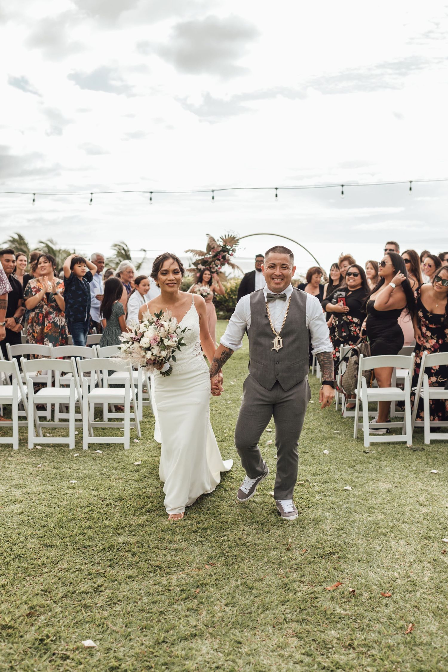 Bride and groom walk down the aisle after outdoor wedding ceremony on green lawn with white chairs and string lights.