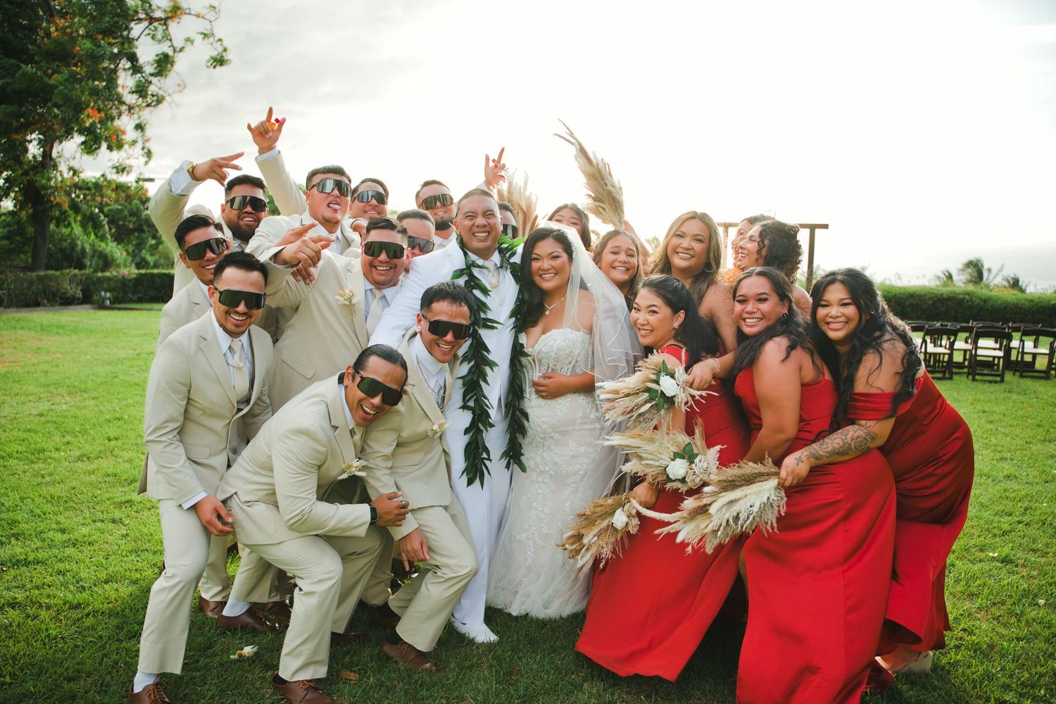 Wedding party celebrating outdoors, groomsmen in tan suits, bridesmaids in red dresses, bride and groom centered on green lawn.