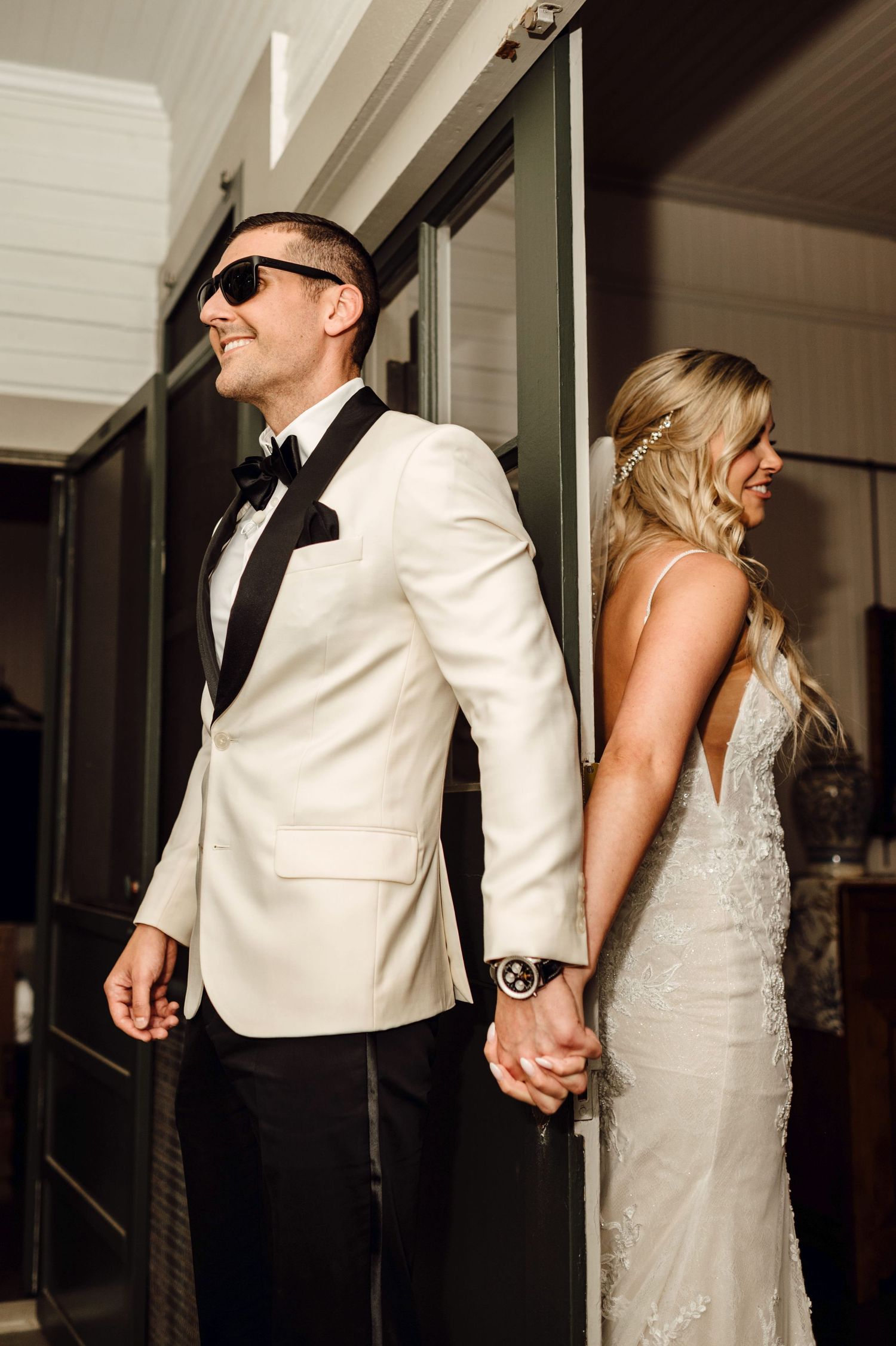 Bride and groom holding hands back-to-back, groom in white tuxedo with sunglasses, bride in lace wedding gown.