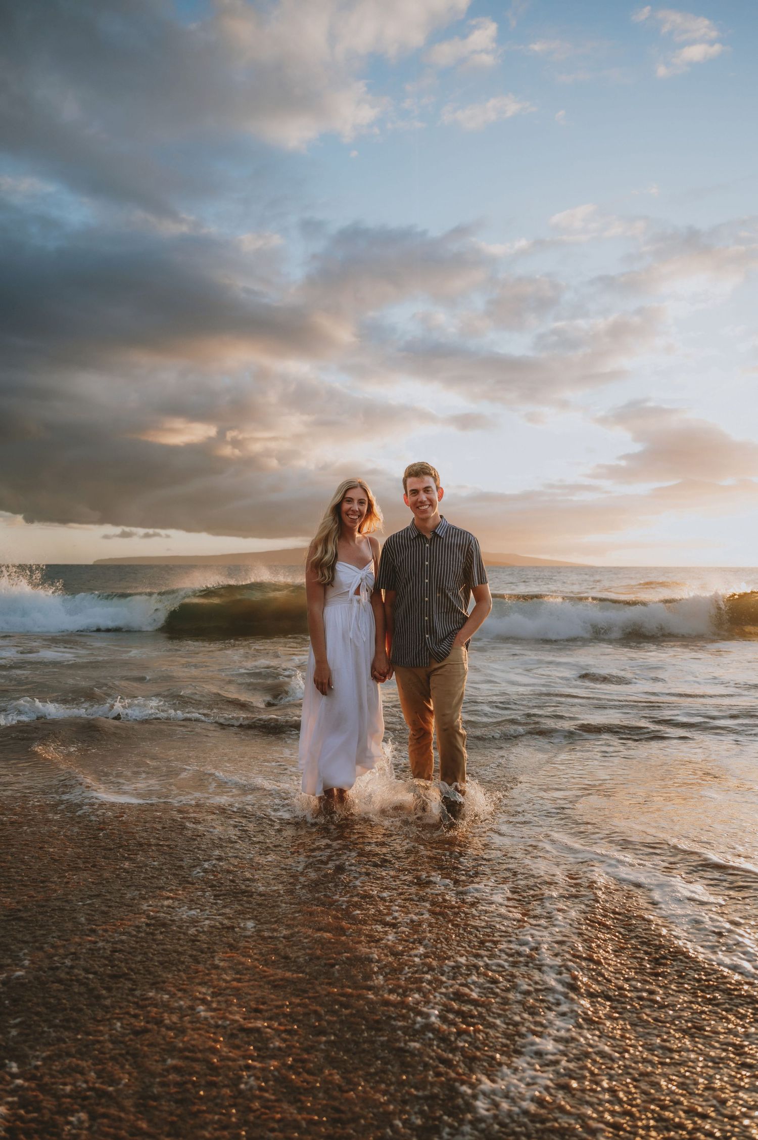 Couple standing in ocean waves at sunset on a sandy beach, woman in white dress, man in gray shirt, golden sky.