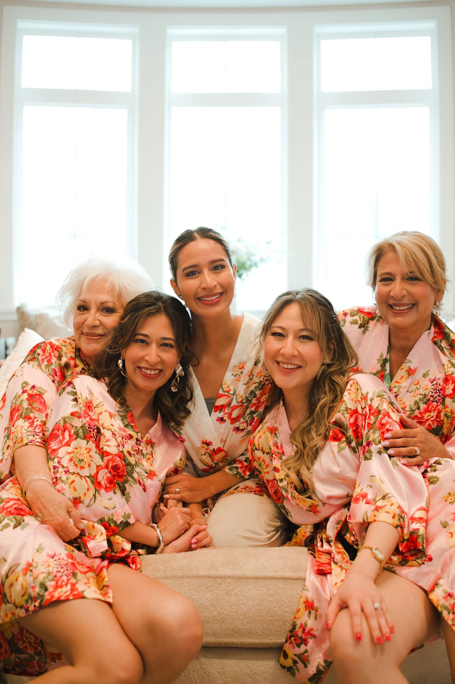 Bride and bridesmaids wearing matching floral robes smile together on a couch during wedding morning preparations.