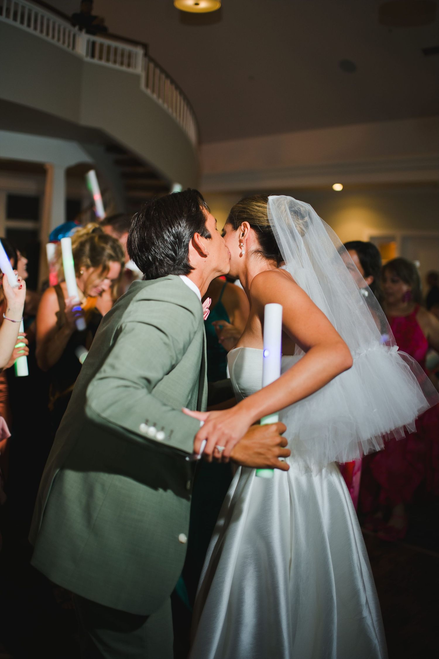 Bride and groom share a romantic kiss at their wedding reception surrounded by guests holding glow sticks.