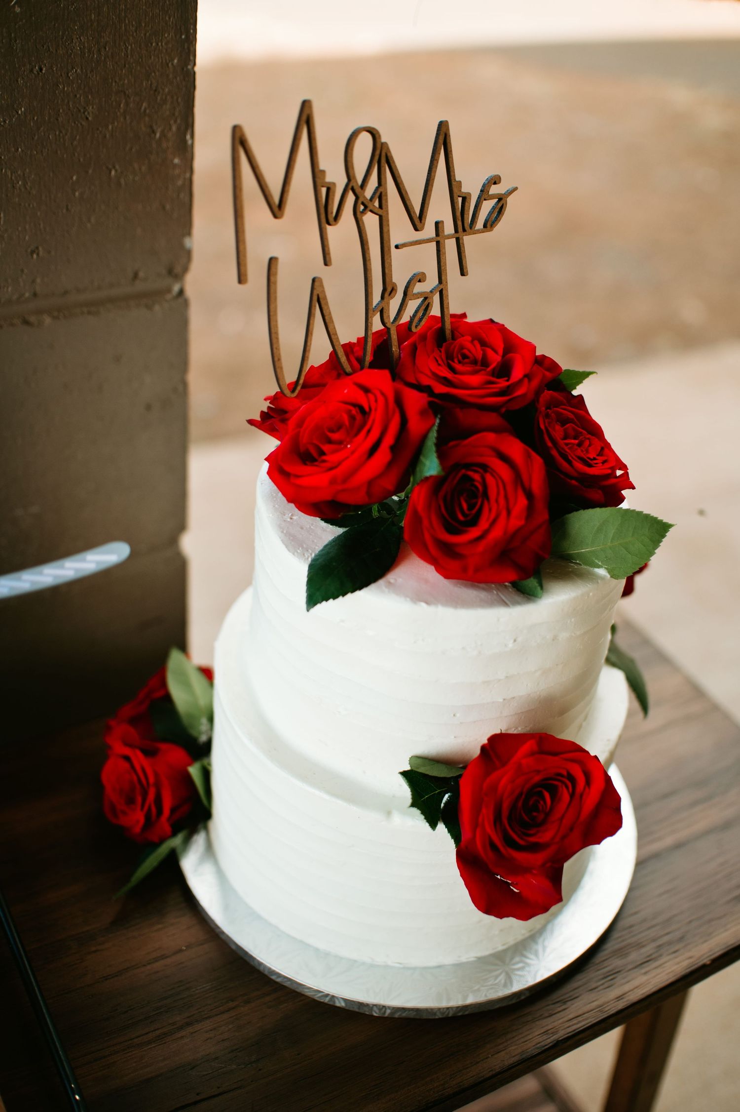Two-tier white wedding cake with red roses and Mr. & Mrs. cake topper on wooden table.