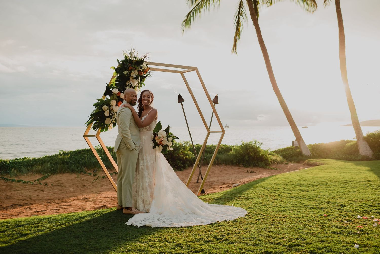 Bride and groom pose under geometric hexagon arch with tropical florals and palm trees at oceanfront wedding.