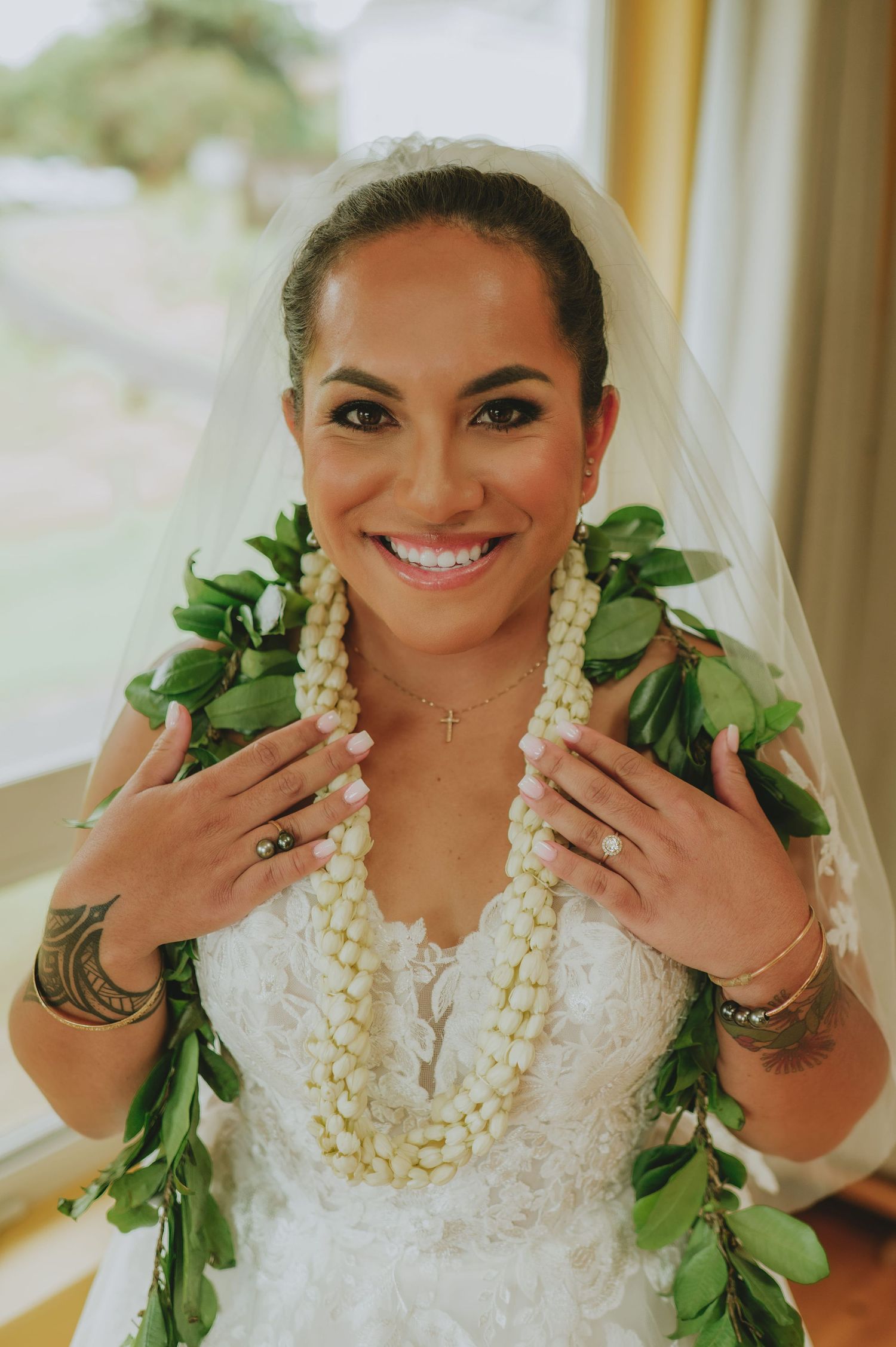 Smiling bride in white lace gown wearing traditional Hawaiian lei and veil on her wedding day.
