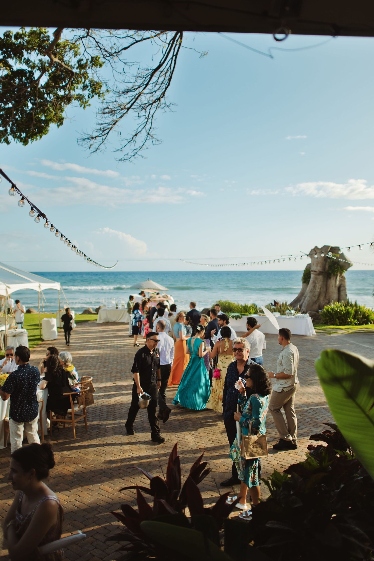 Guests mingle at an oceanfront wedding reception venue with a rocky coastline and blue sky backdrop.