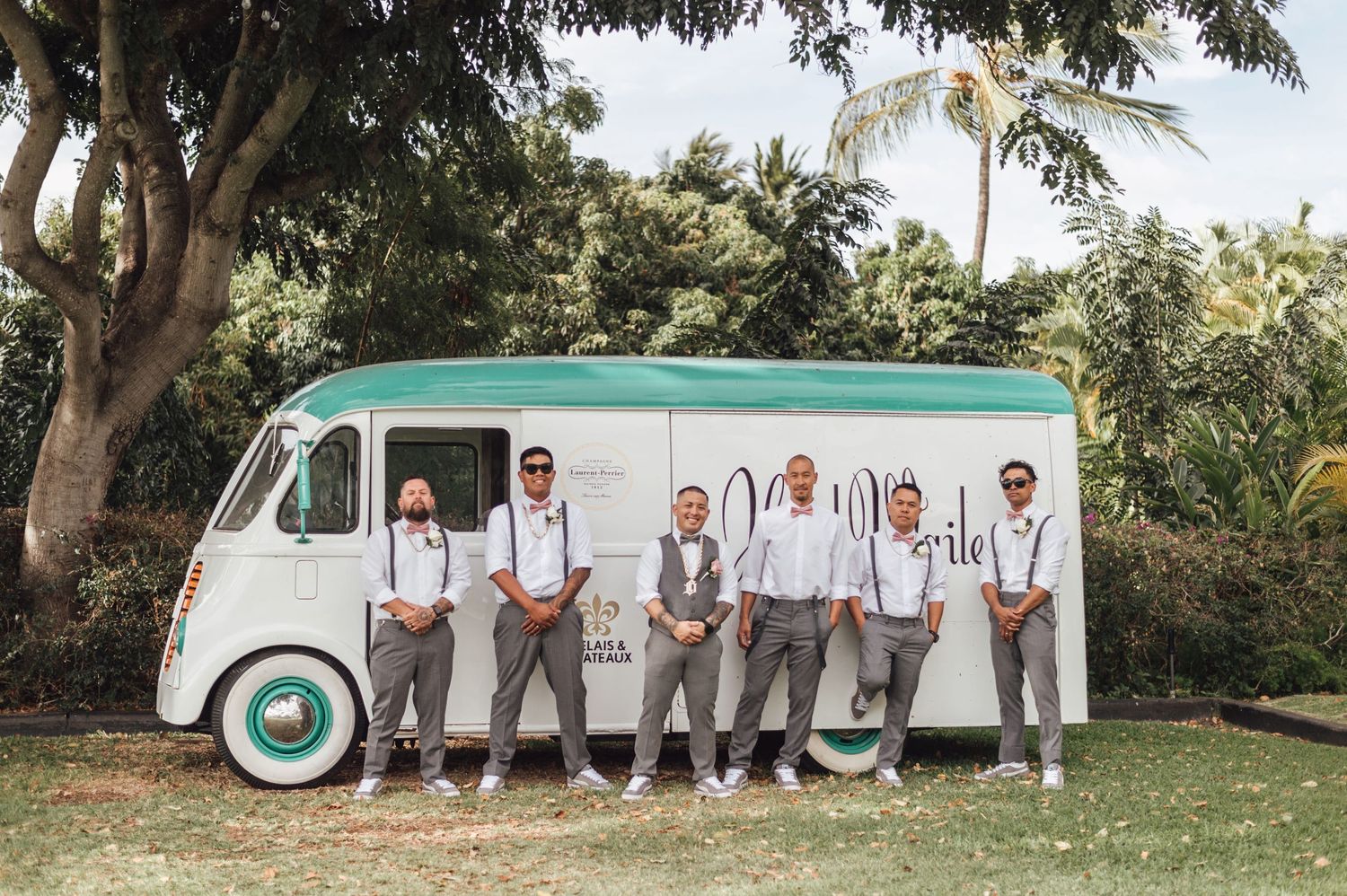 Groomsmen in gray suits with suspenders posing in front of a vintage mint green food truck at an outdoor wedding.