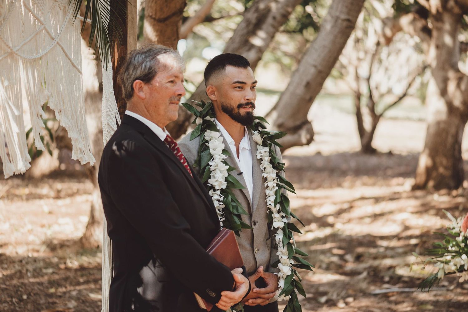 Groom wearing floral lei stands with officiant during outdoor wedding ceremony among trees.