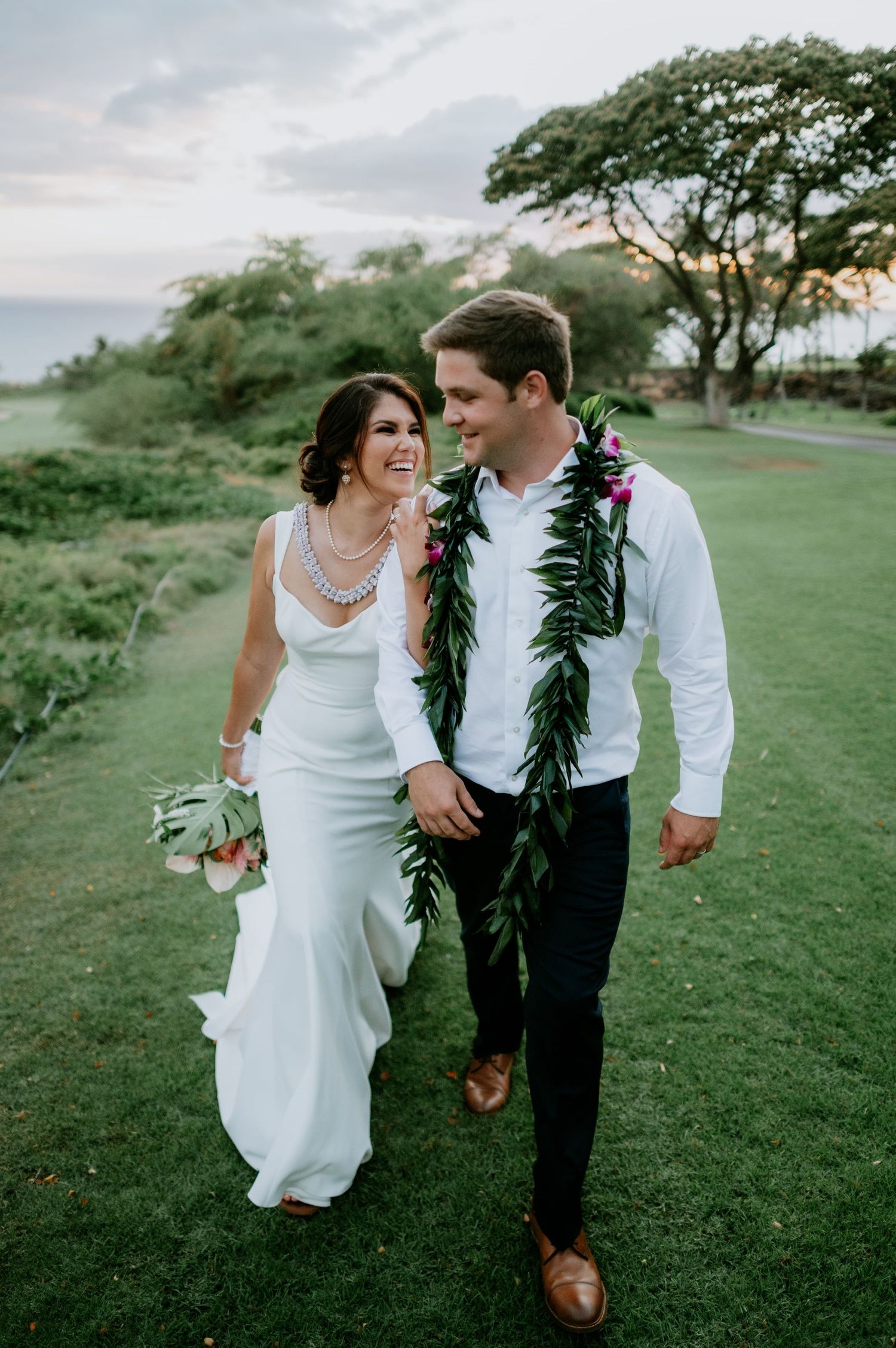 Newlyweds walk and laugh together on a grassy oceanside path.