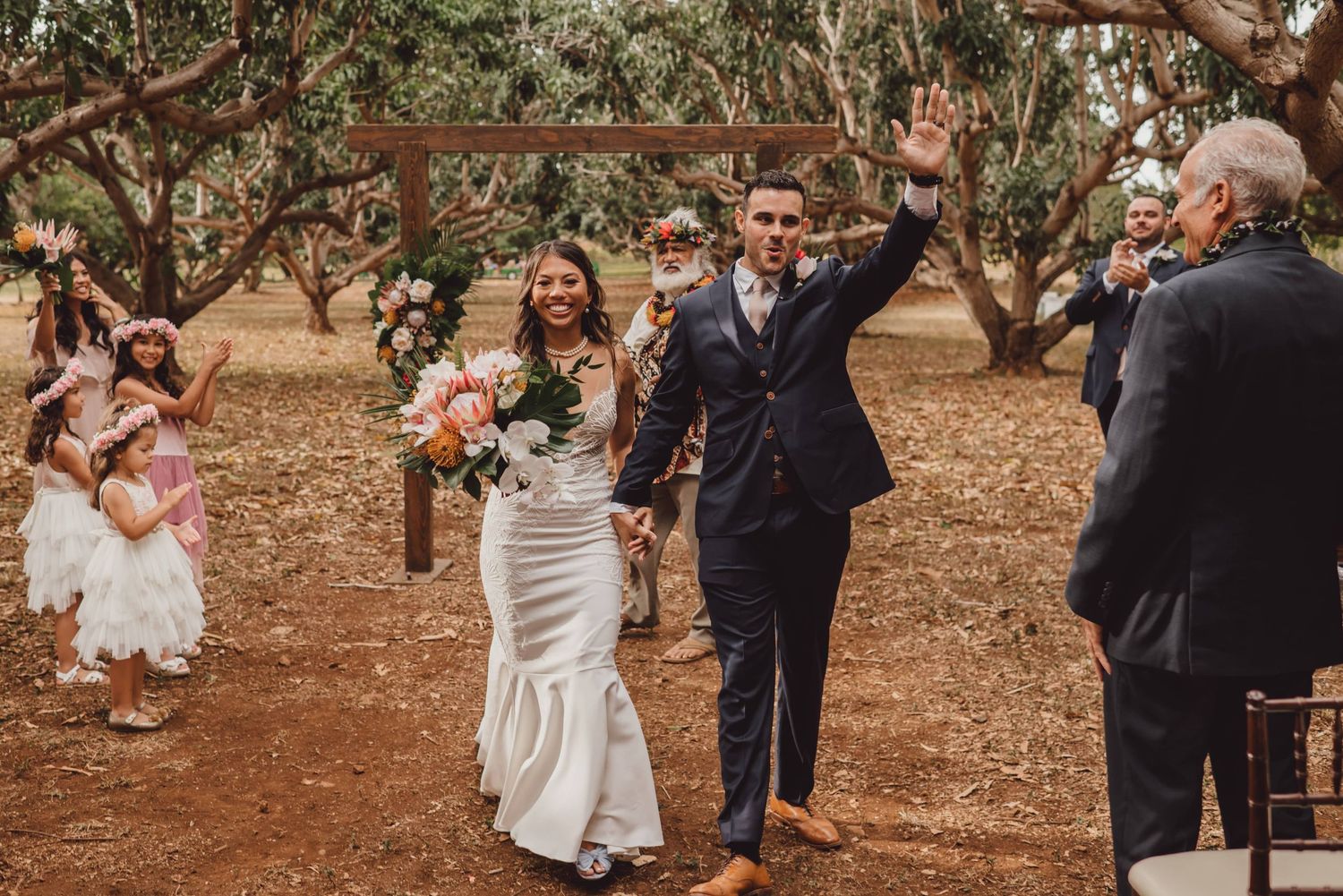 Romantic wedding couple embraces under rustic trees in an outdoor garden setting.