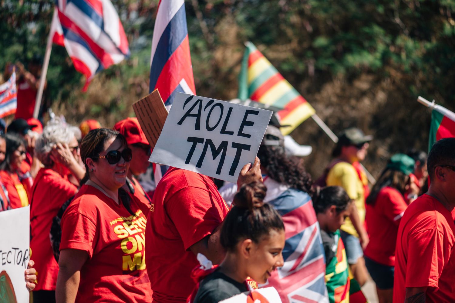 Demonstrators in red shirts hold protest signs and Hawaiian flags during a rally.