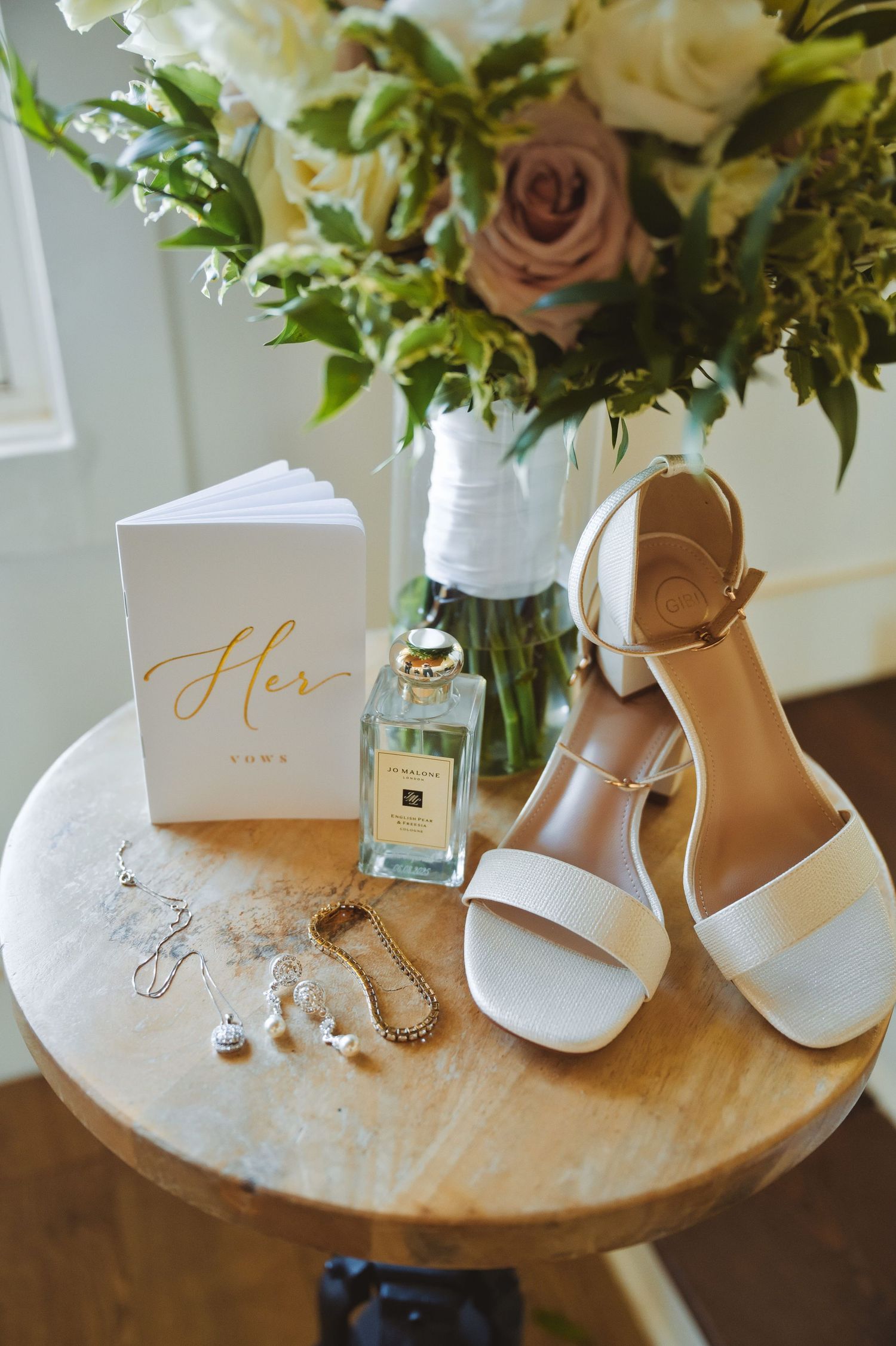 Wedding flat lay with white heels, Jo Malone perfume, earrings, vow book, and floral bouquet.