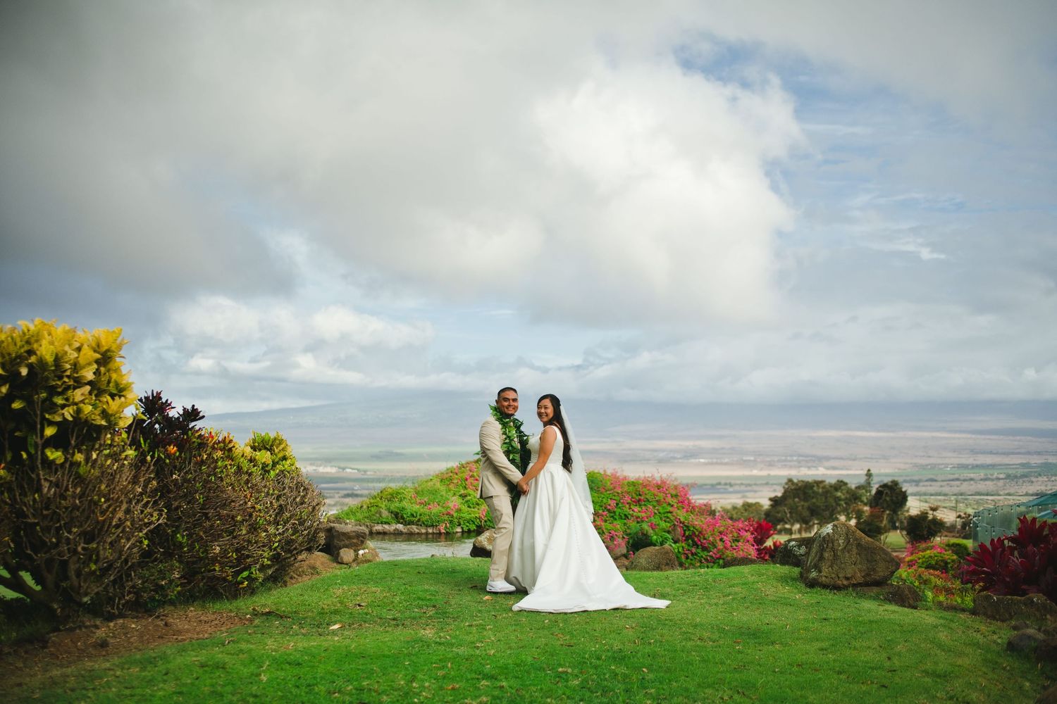 Bride and groom pose on lush green hillside with panoramic ocean views and colorful flowers under dramatic cloudy sky.