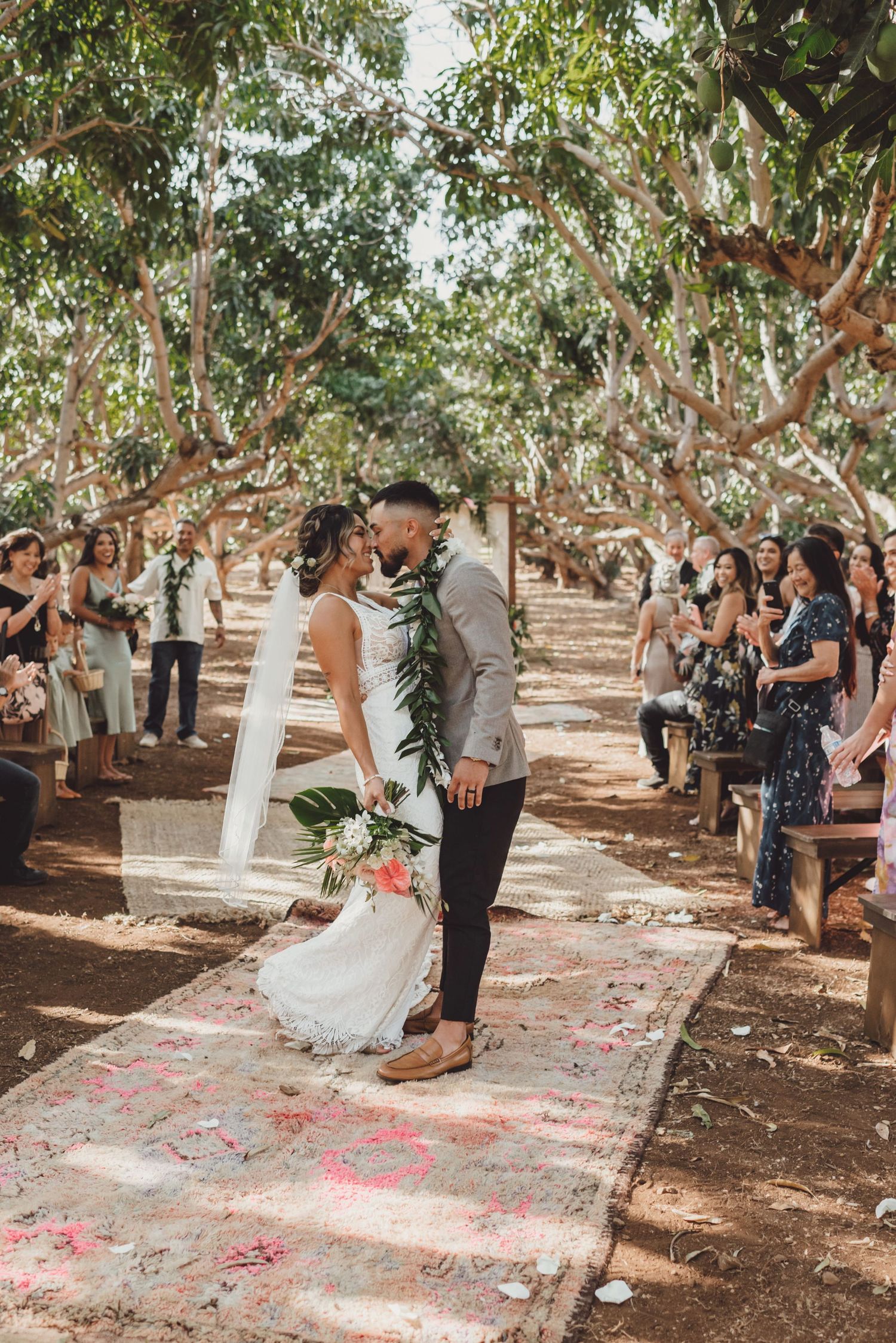 Bride and groom share first kiss at outdoor wedding ceremony in tropical tree grove with guests seated on wooden benches.