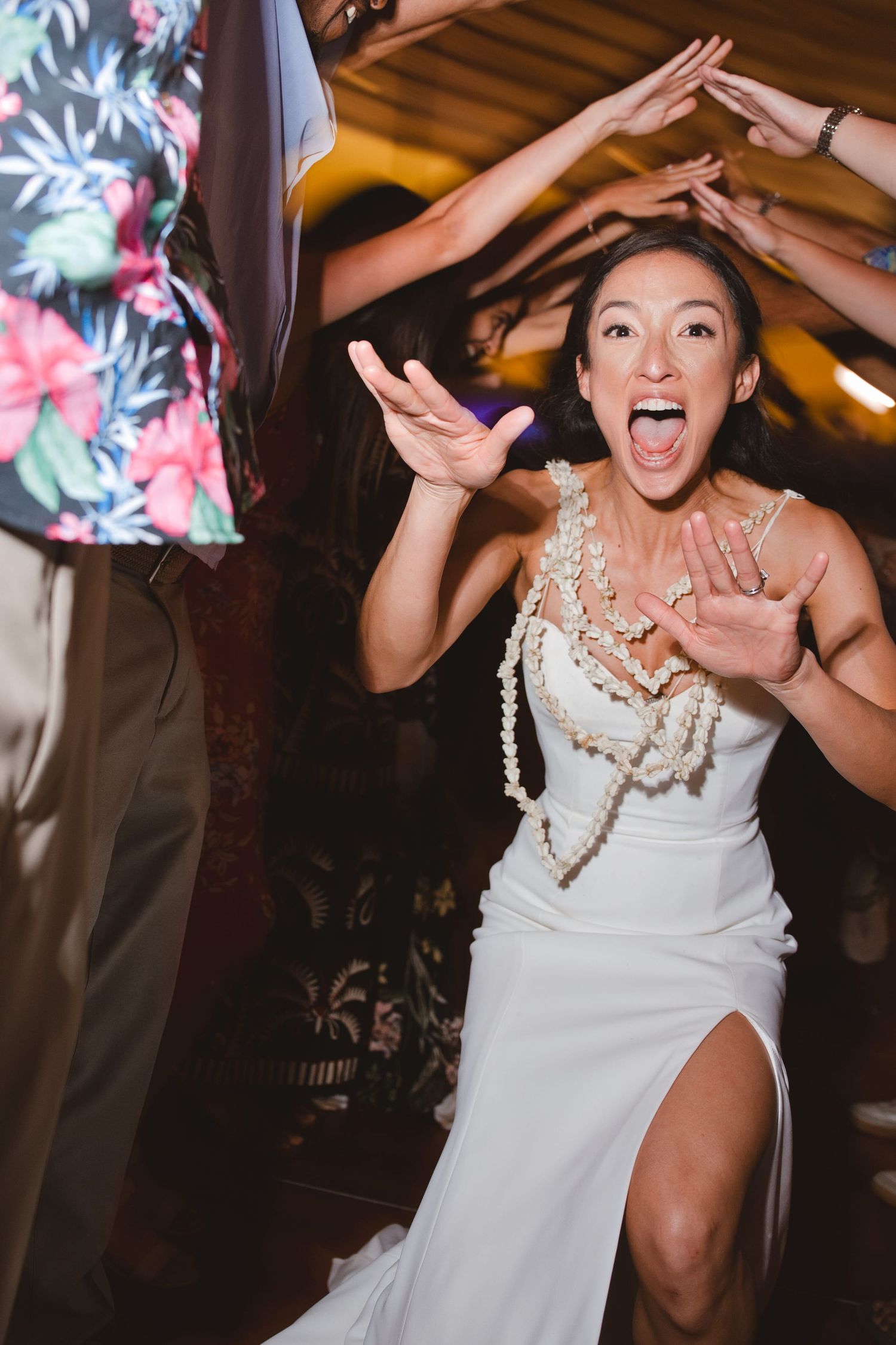 Bride in white gown with pearl necklaces dancing joyfully with hands raised at wedding reception celebration.