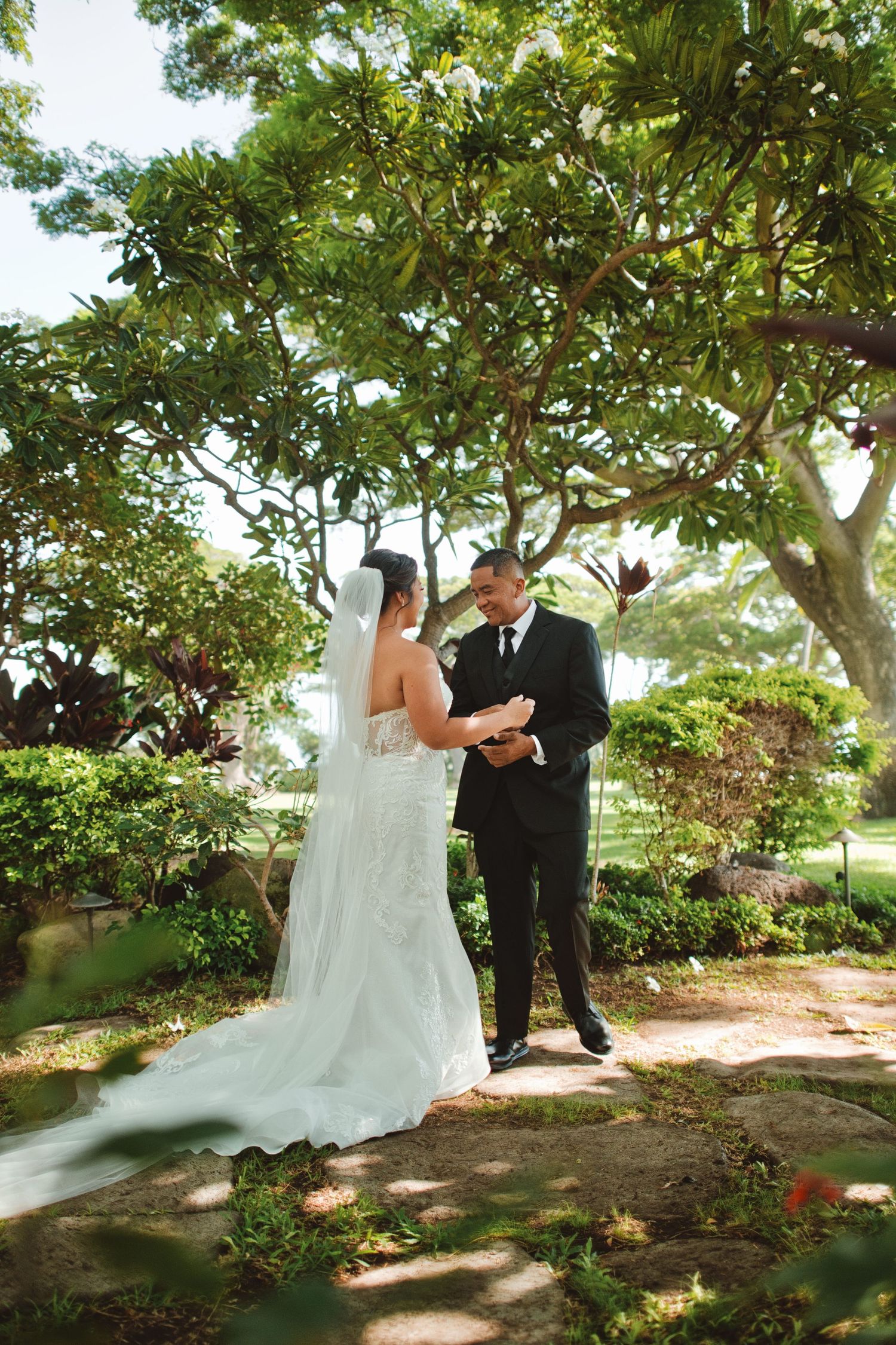 Bride and groom share a tender first look moment on a garden path surrounded by lush tropical trees and greenery.