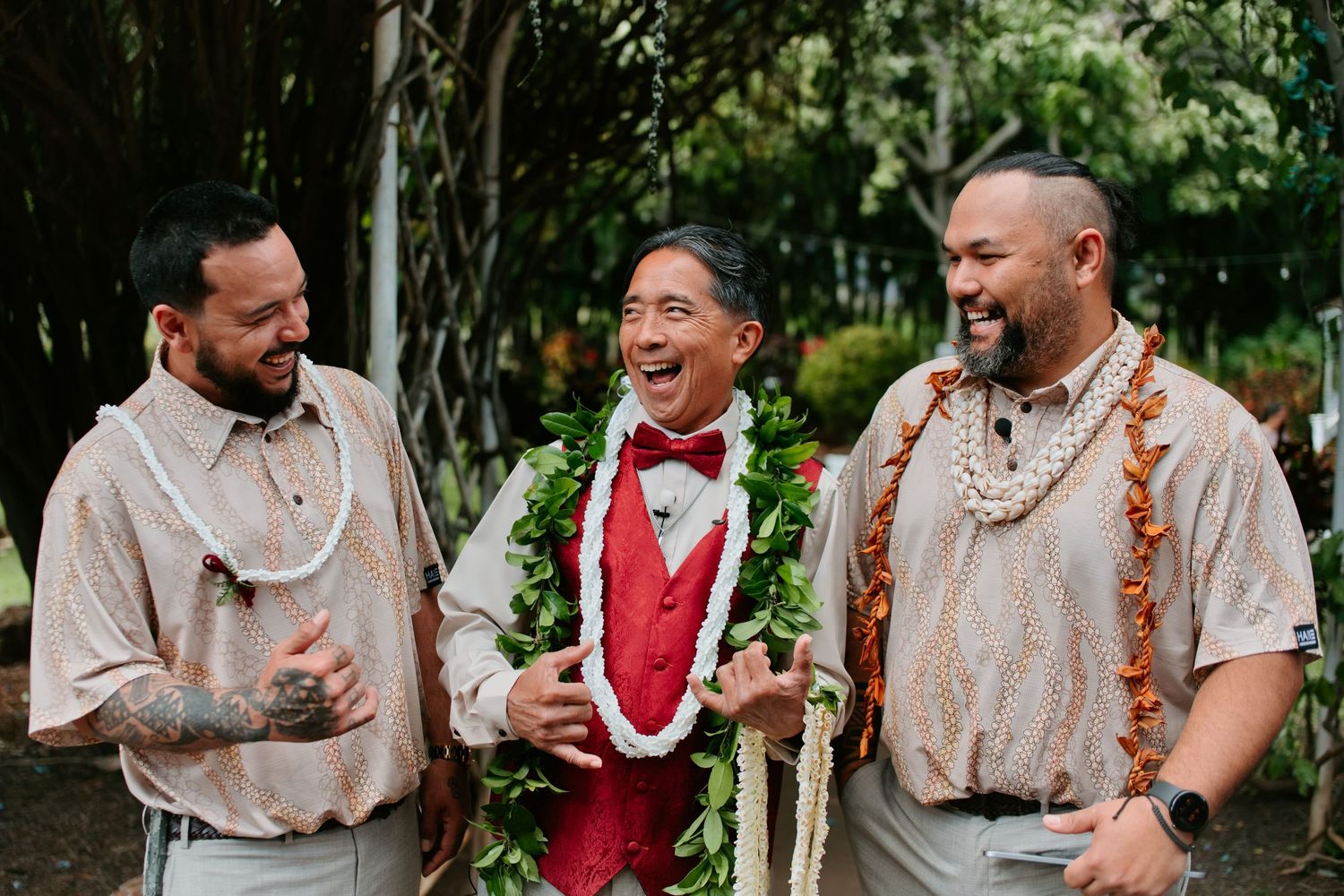 Three men laughing at a Hawaiian wedding, groom in red vest wearing lei, groomsmen in beige shirts with floral leis.