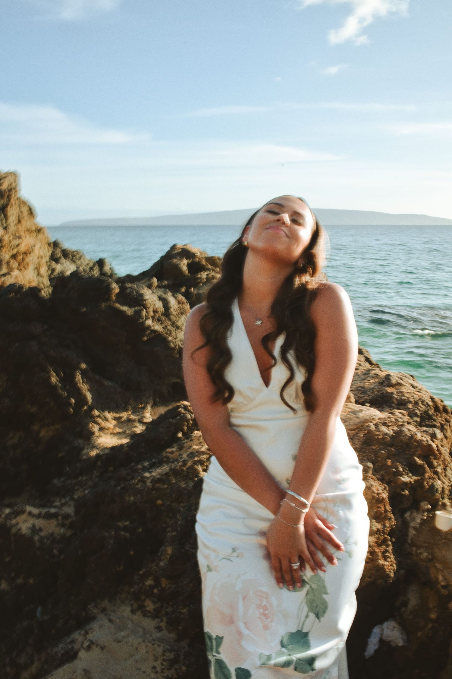 Woman in floral white dress posing on rocky beach with ocean backdrop in Hawaii.
