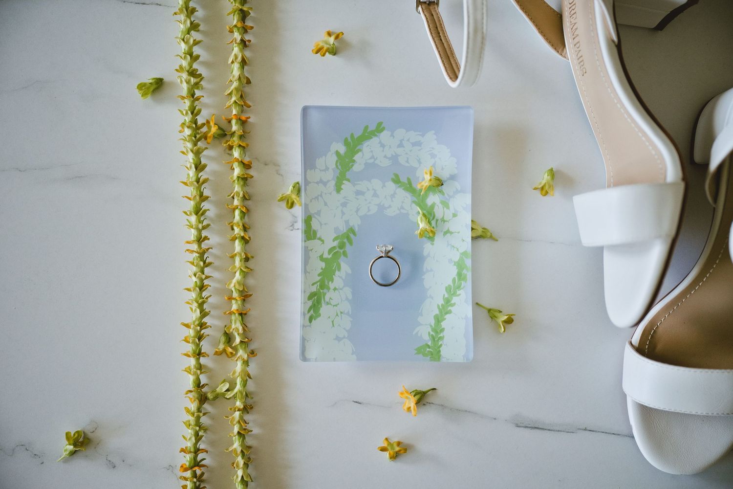 Wedding ring resting on floral illustrated card beside white heels and yellow flower garlands.