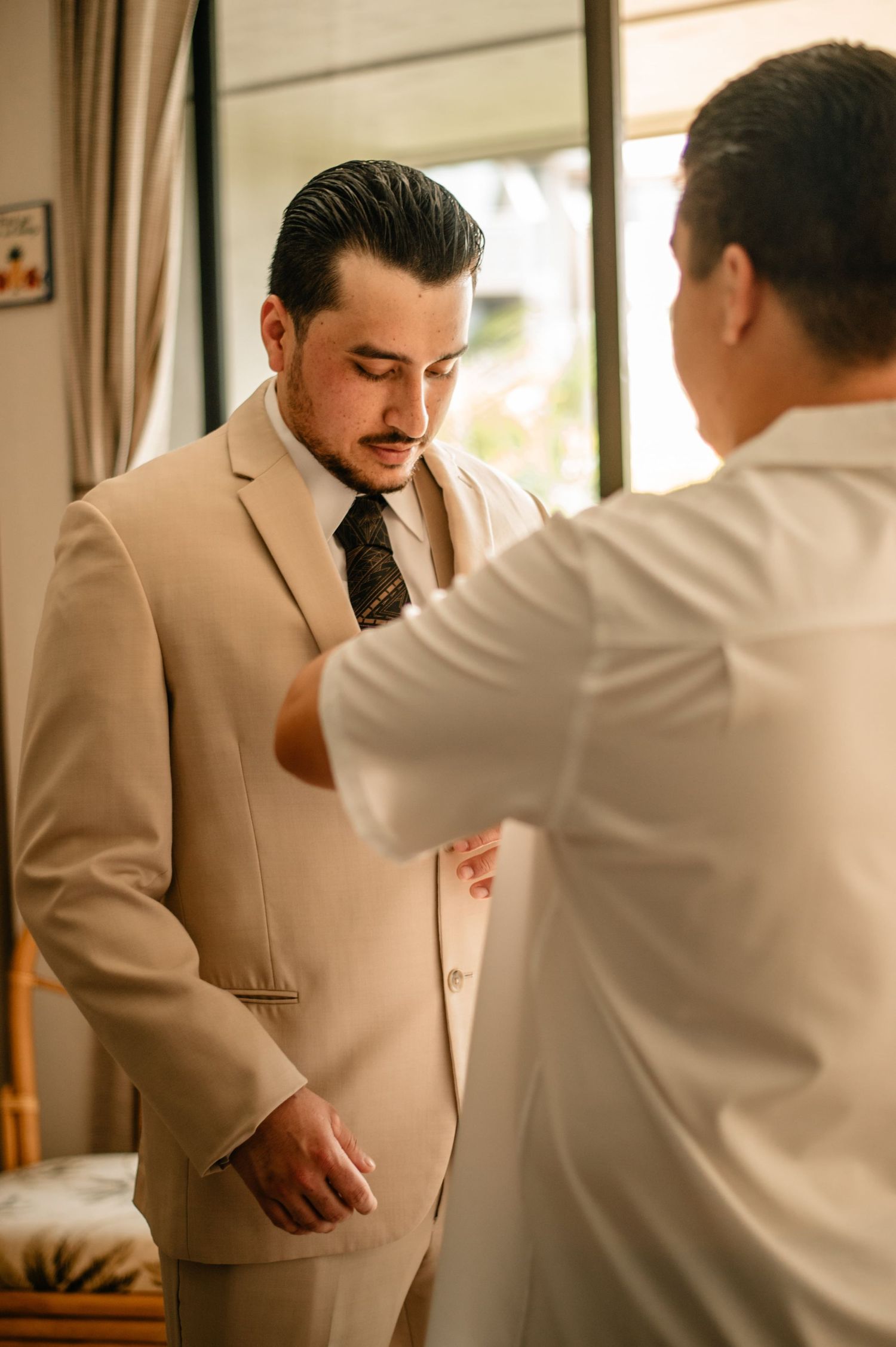 Groom getting dressed in a beige suit with help from others in a sunlit room.