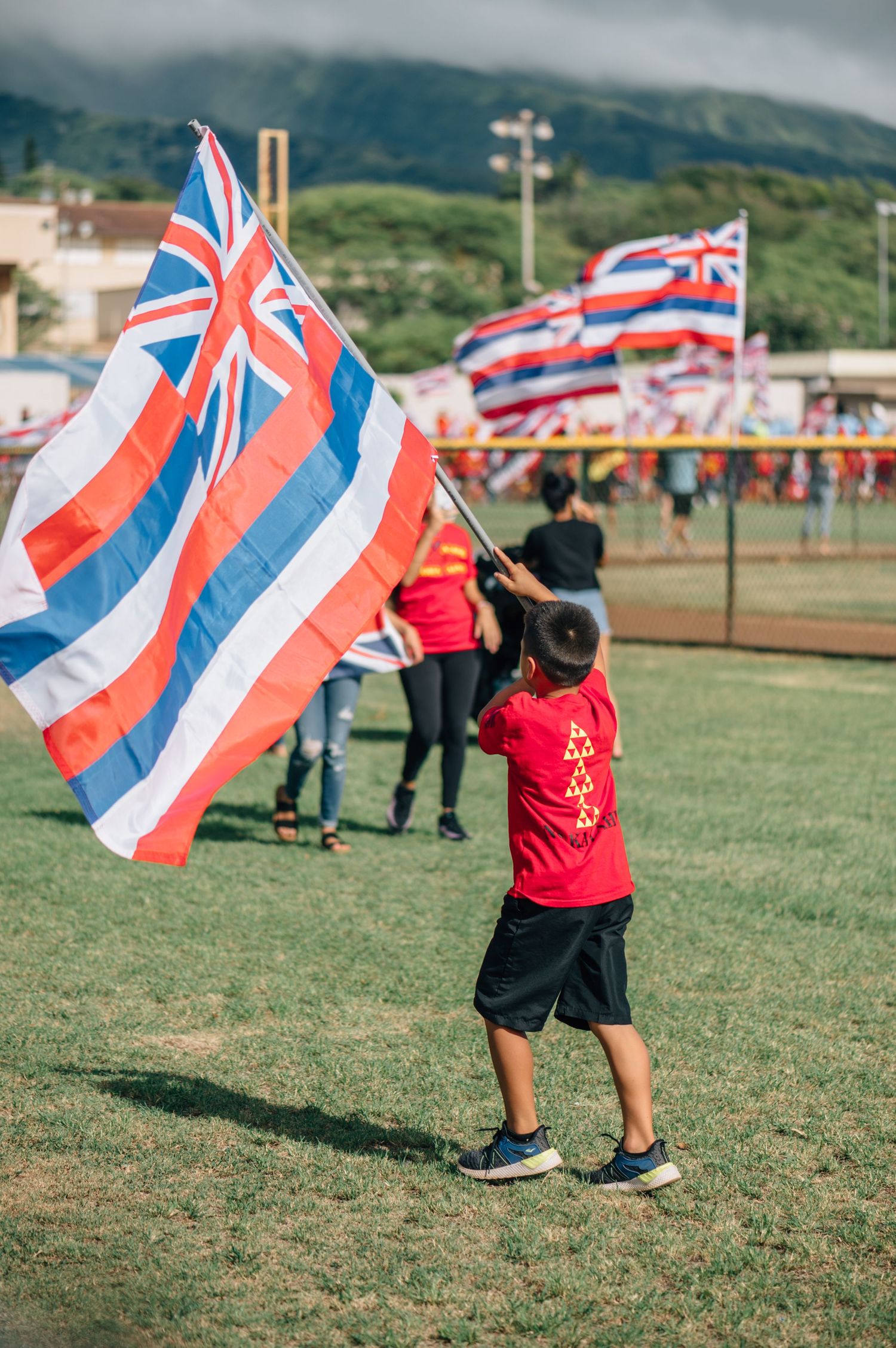 Children wave Hawaiian state flags on a grassy field with mountains behind them.