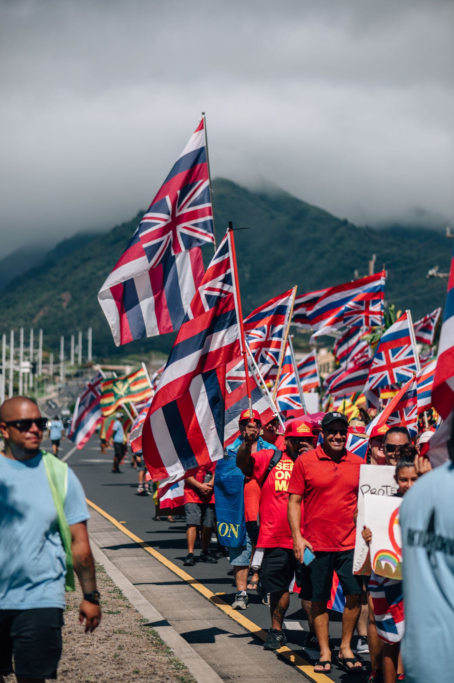 Large crowd gathers along a coastal road waving Hawaiian flags with mountains in the background.