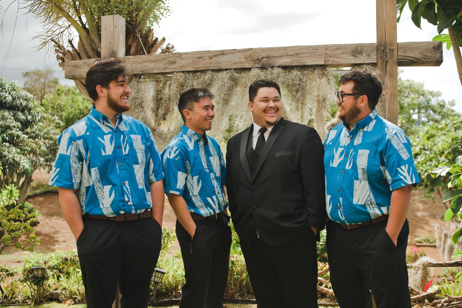 Groom in black suit stands with three groomsmen wearing matching blue Hawaiian shirts at an outdoor wedding.
