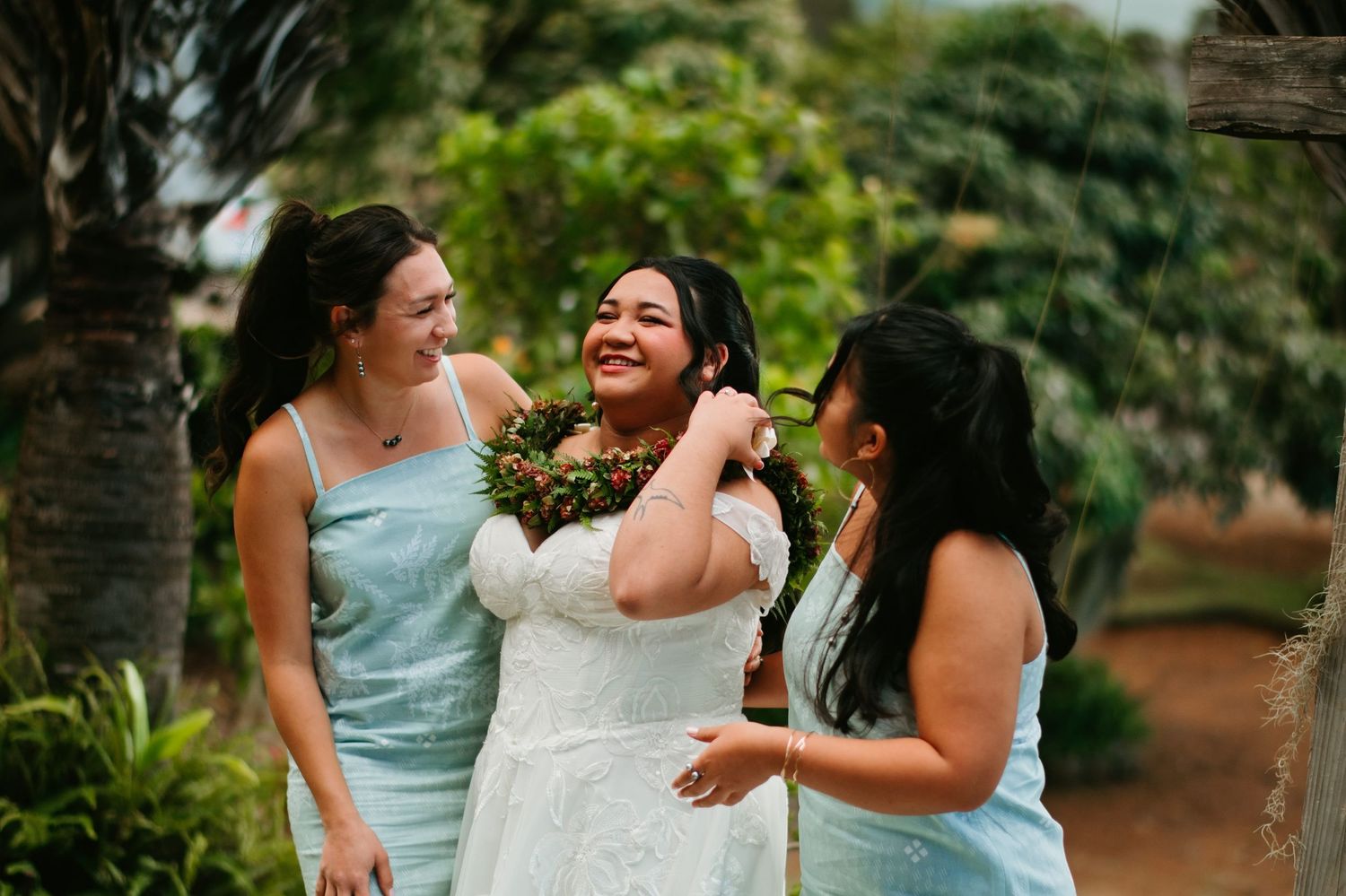 Bride in white dress laughs with two bridesmaids in light blue dresses outdoors at a tropical wedding.