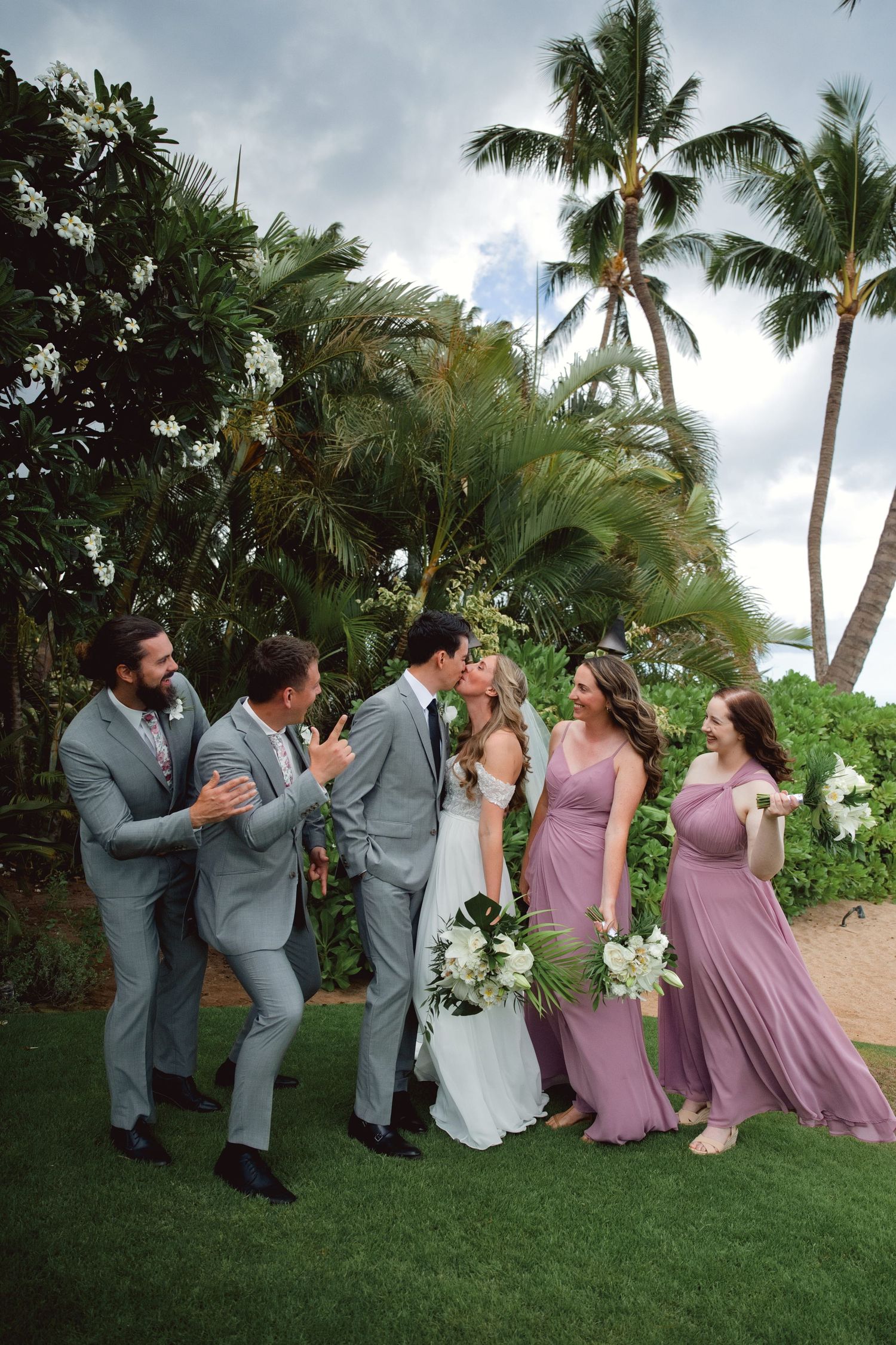 Wedding party kissing and celebrating outdoors in Hawaii, groomsmen in gray suits and bridesmaids in mauve dresses with tropical palms.